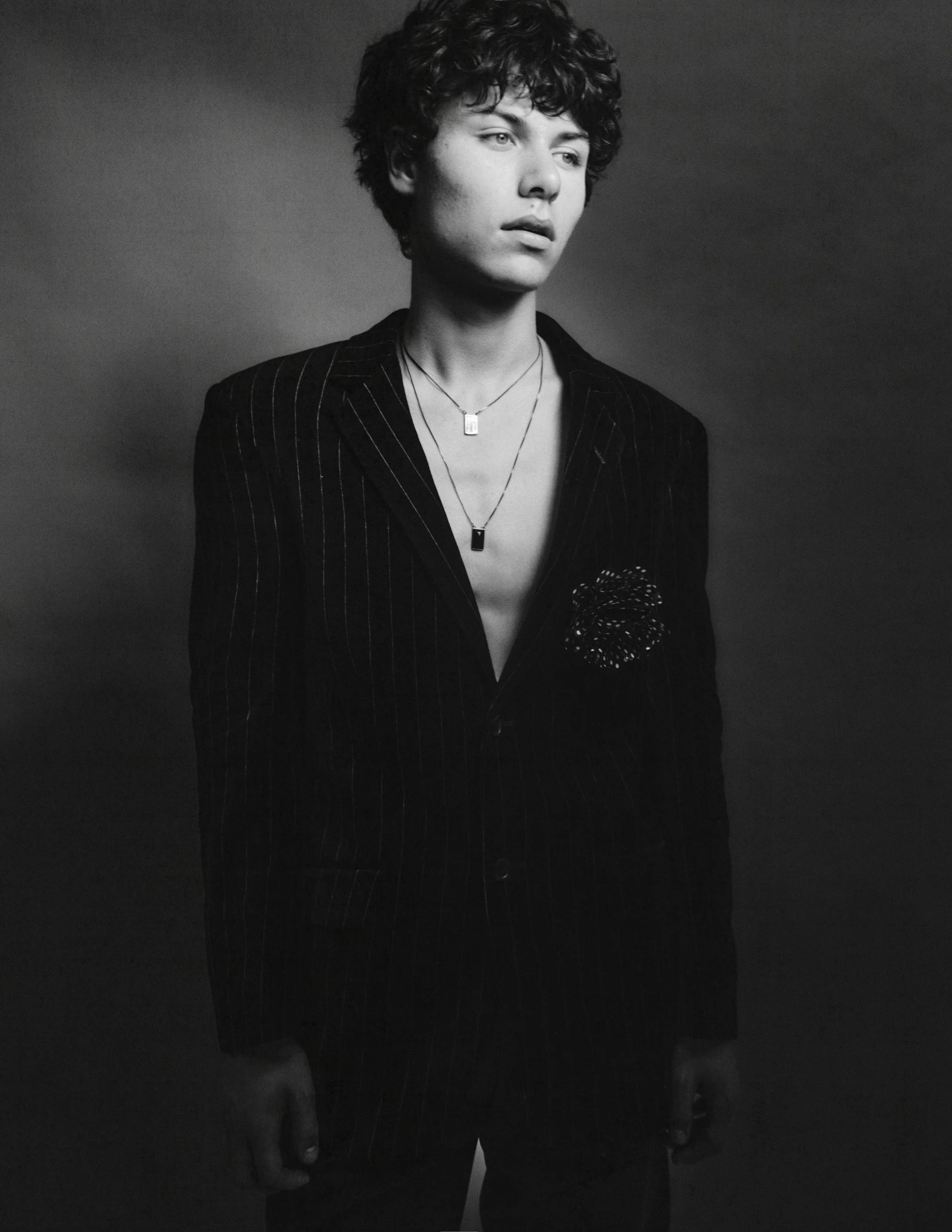 Black and white portrait of a young man with curly hair, wearing a pinstripe blazer with a floral embellishment, layered necklaces, and an open shirt.