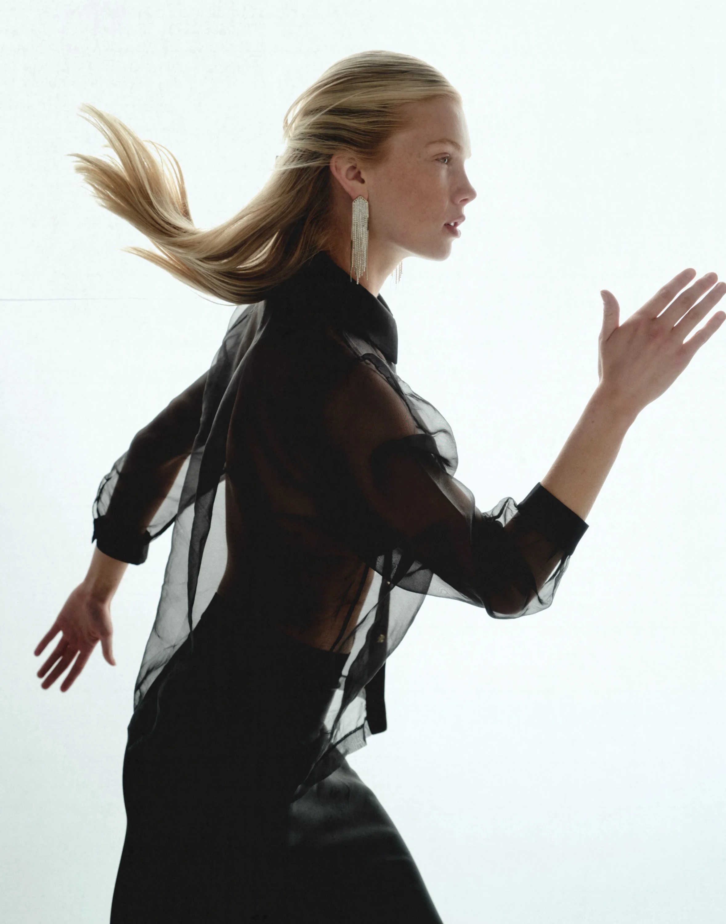 A woman with blonde hair running in profile, wearing a sheer black blouse and long earrings, against a light background.