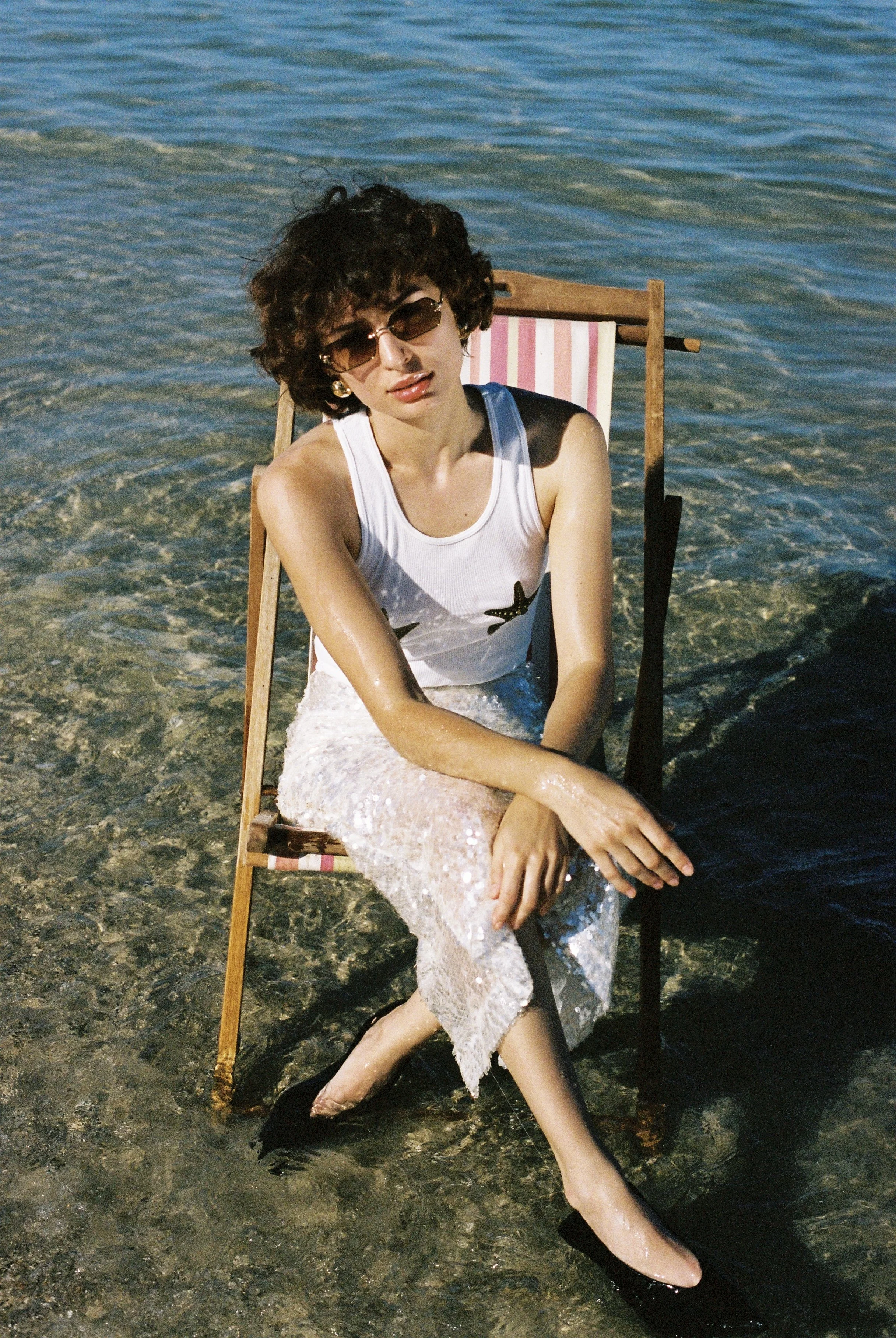 Woman sitting on a beach chair in shallow water, wearing sunglasses, a white sleeveless top, and a sparkly skirt, with one leg crossed over the other and water splashing around her.