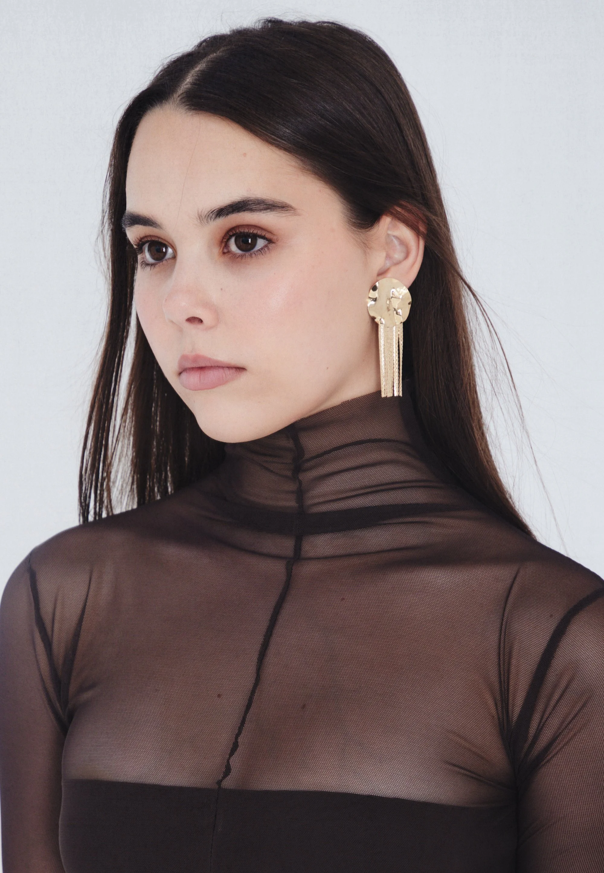 A young woman with long dark hair wears a sheer black top and large gold earrings, looking to her left.