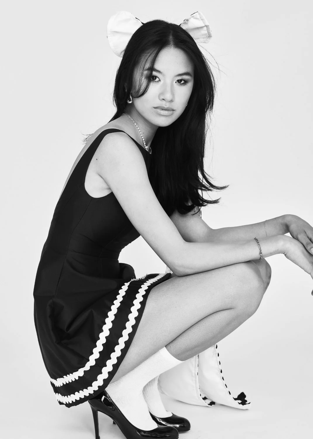 Black and white photo of a young woman squatting, wearing black high heels, white socks, a black dress with white trim, and a bow headband. She has long dark hair and is looking into the camera.