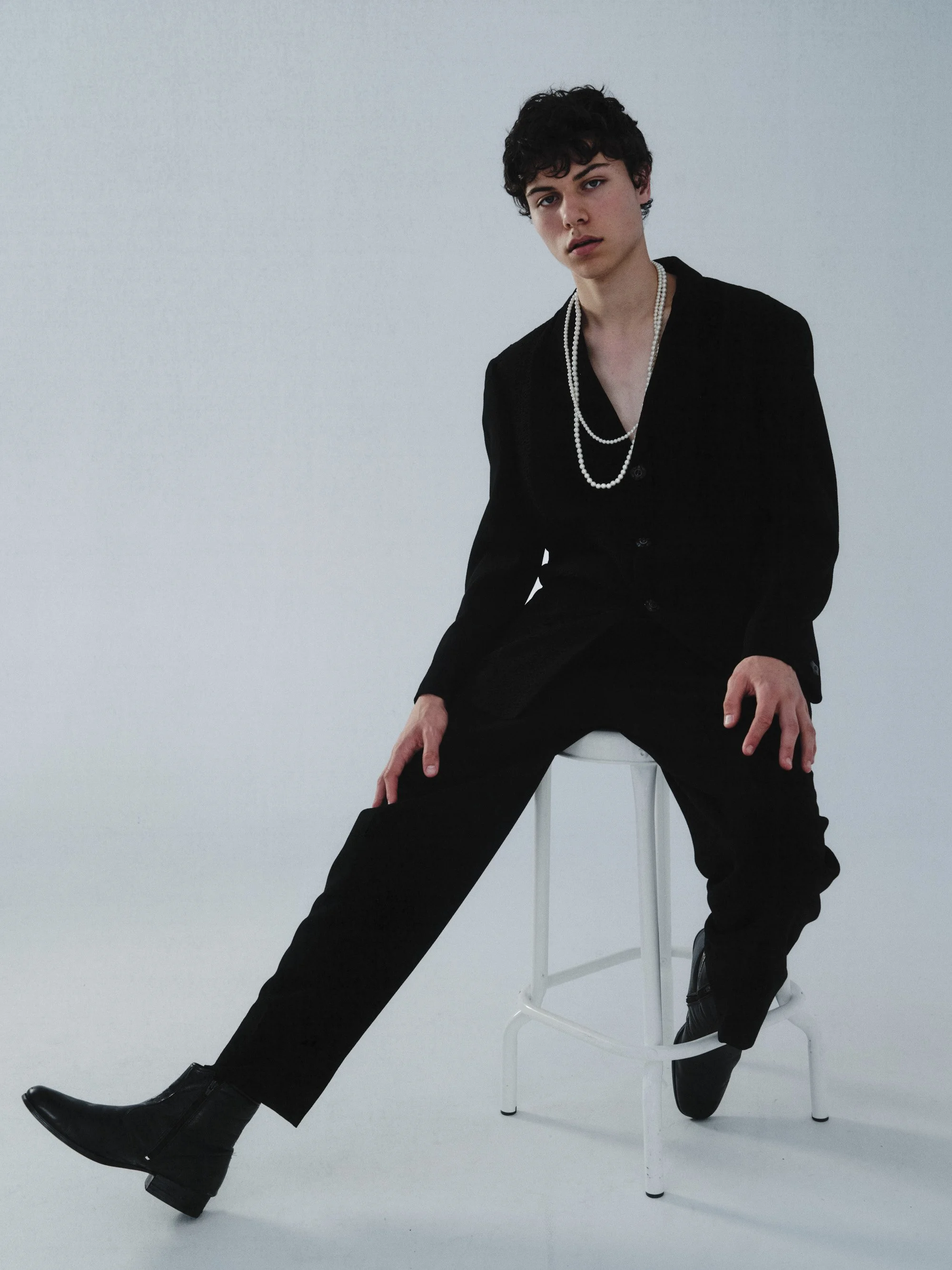 Person with short curly hair wearing a black suit and pearl necklace sitting on a white stool against a plain background.