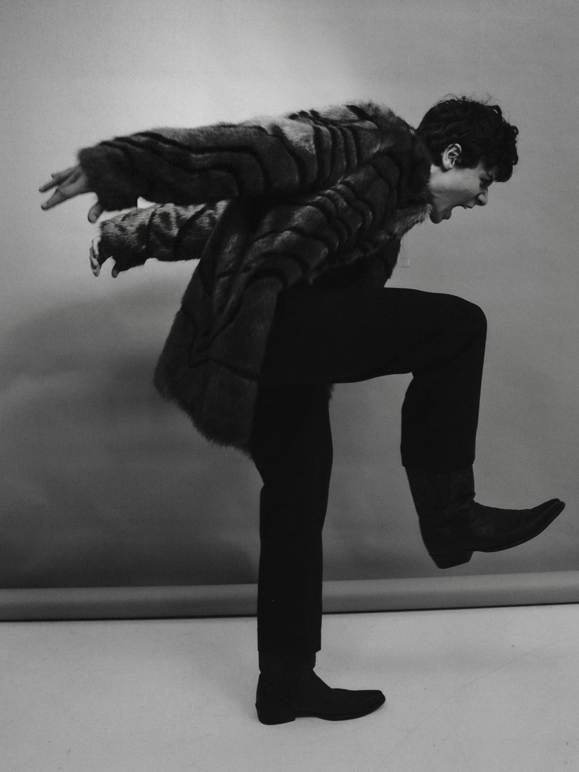 A person in a fur coat striking a dynamic pose, balancing on one leg with the other leg raised, against a plain backdrop.