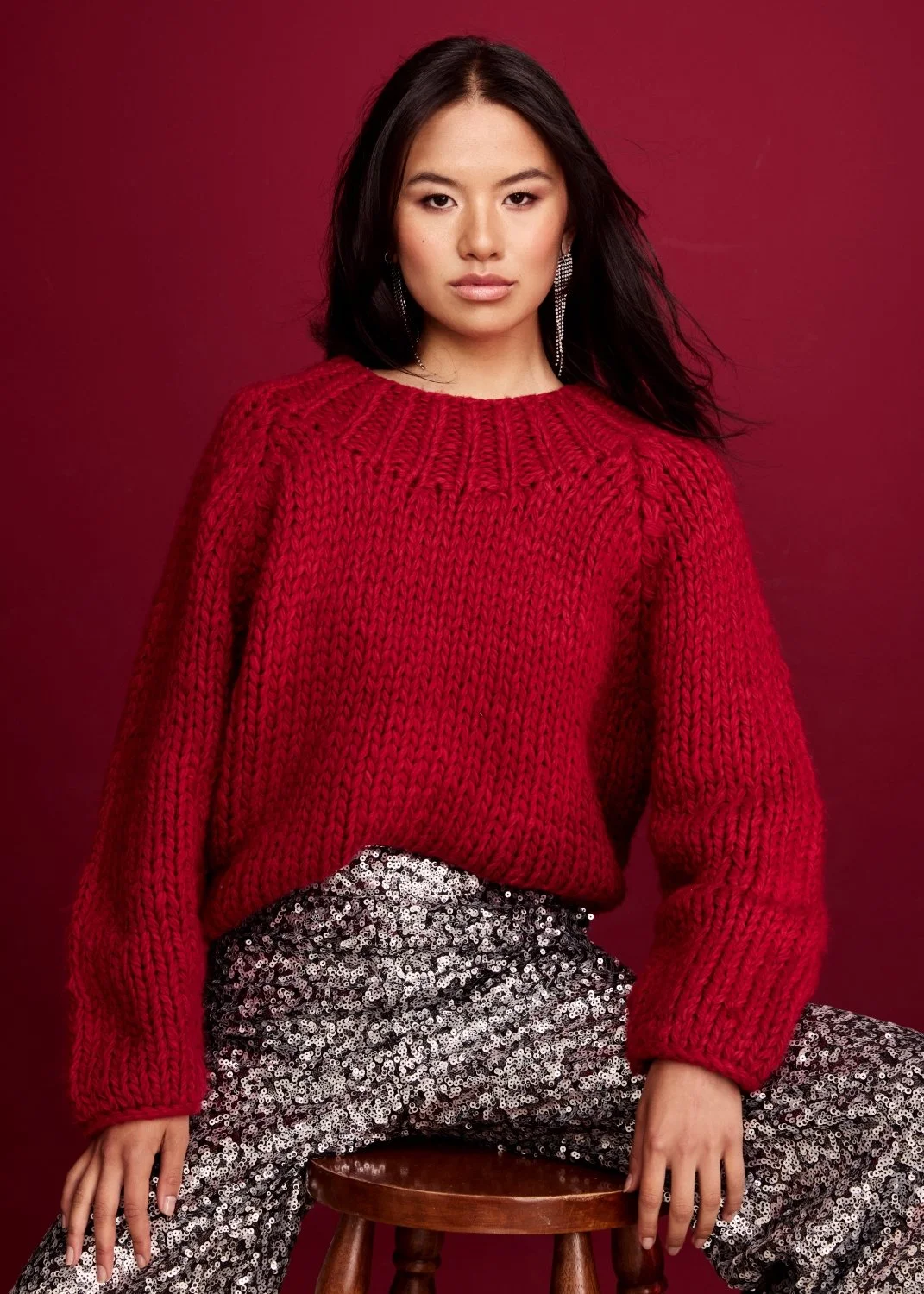 A young woman with dark hair in a red knitted sweater and sequin pants sitting on a wooden stool against a red background.