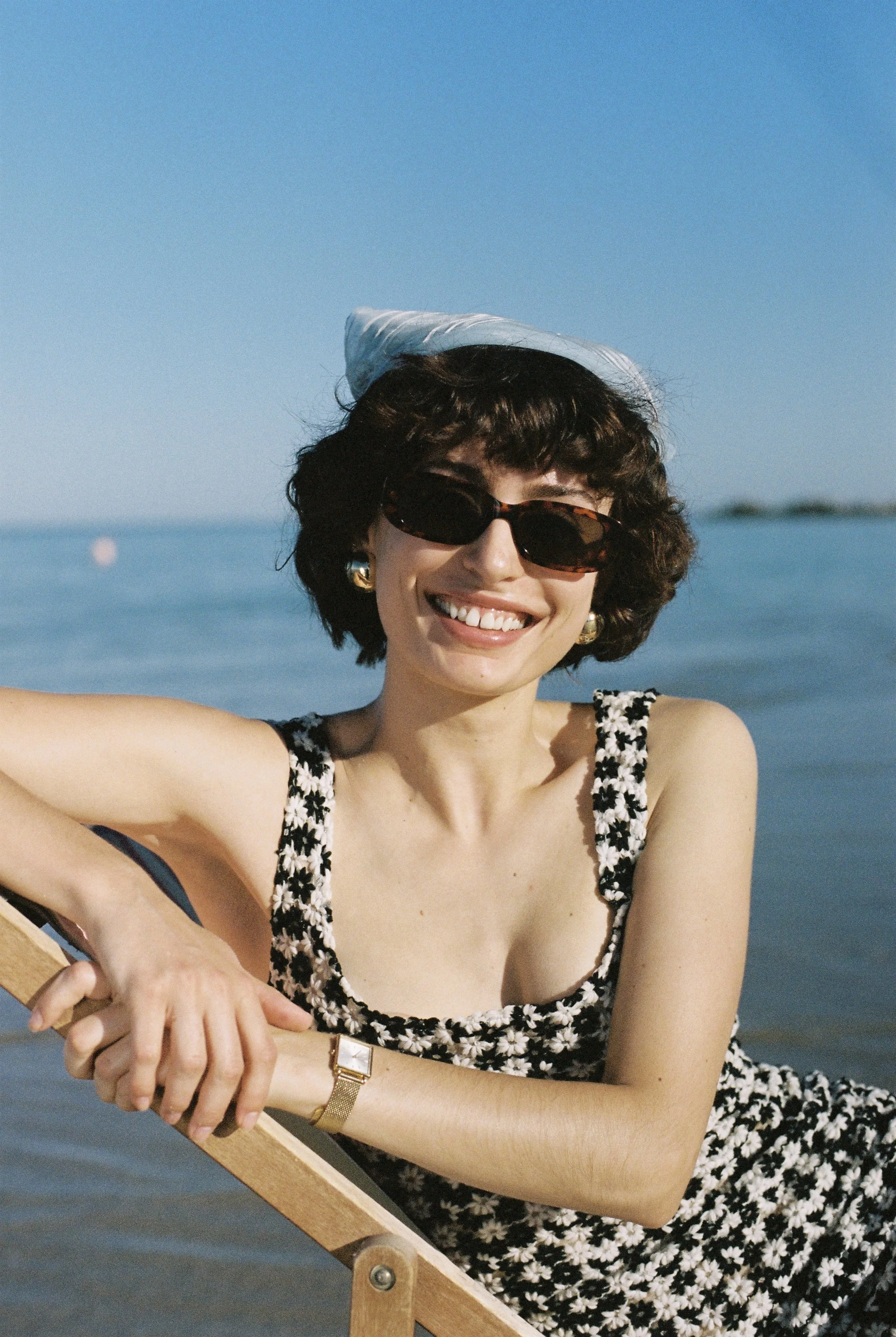 A woman with short dark curly hair wearing sunglasses, a white hat, gold earrings, and a black and white floral dress, smiling while sitting on a deck near the water with a blue sky in the background.