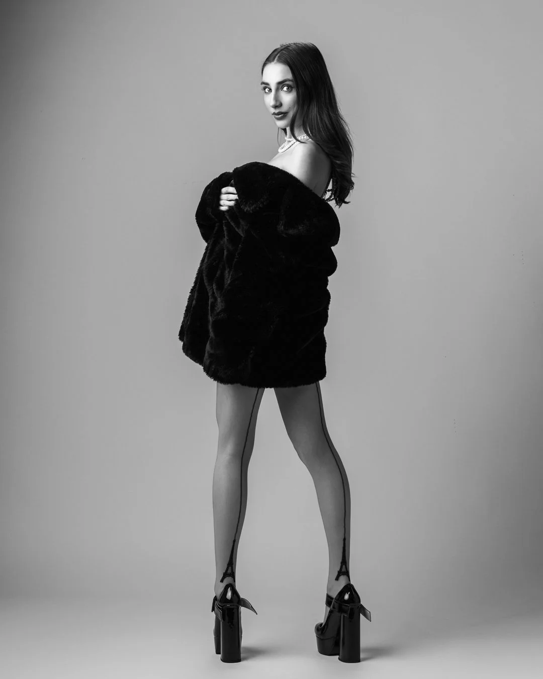 A woman with dark hair in high heels, wearing transparent patterned stockings and a fur coat, looking at the camera against a plain background.