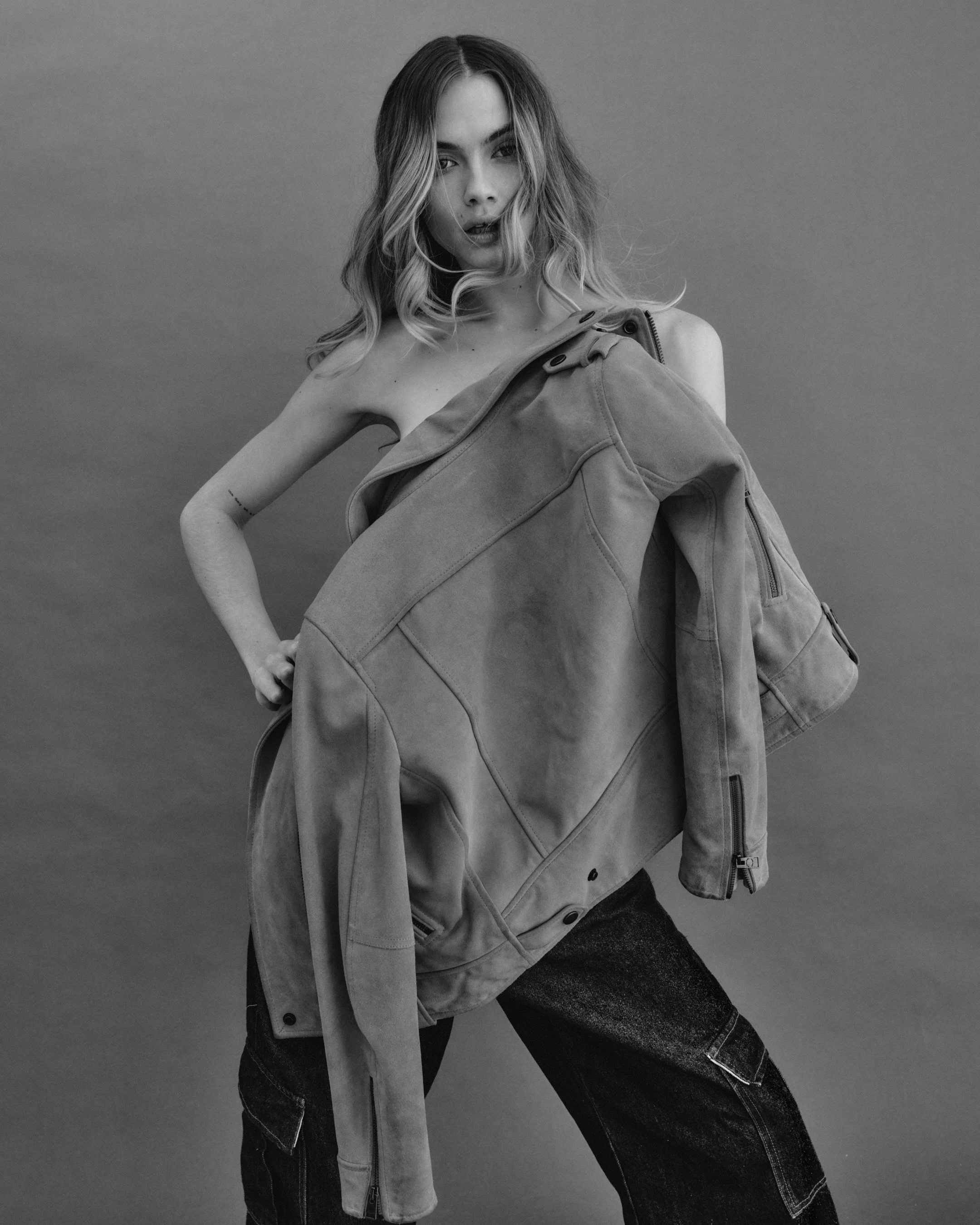 Black and white photo of a woman with wavy hair holding a large jacket, posing against a plain background.