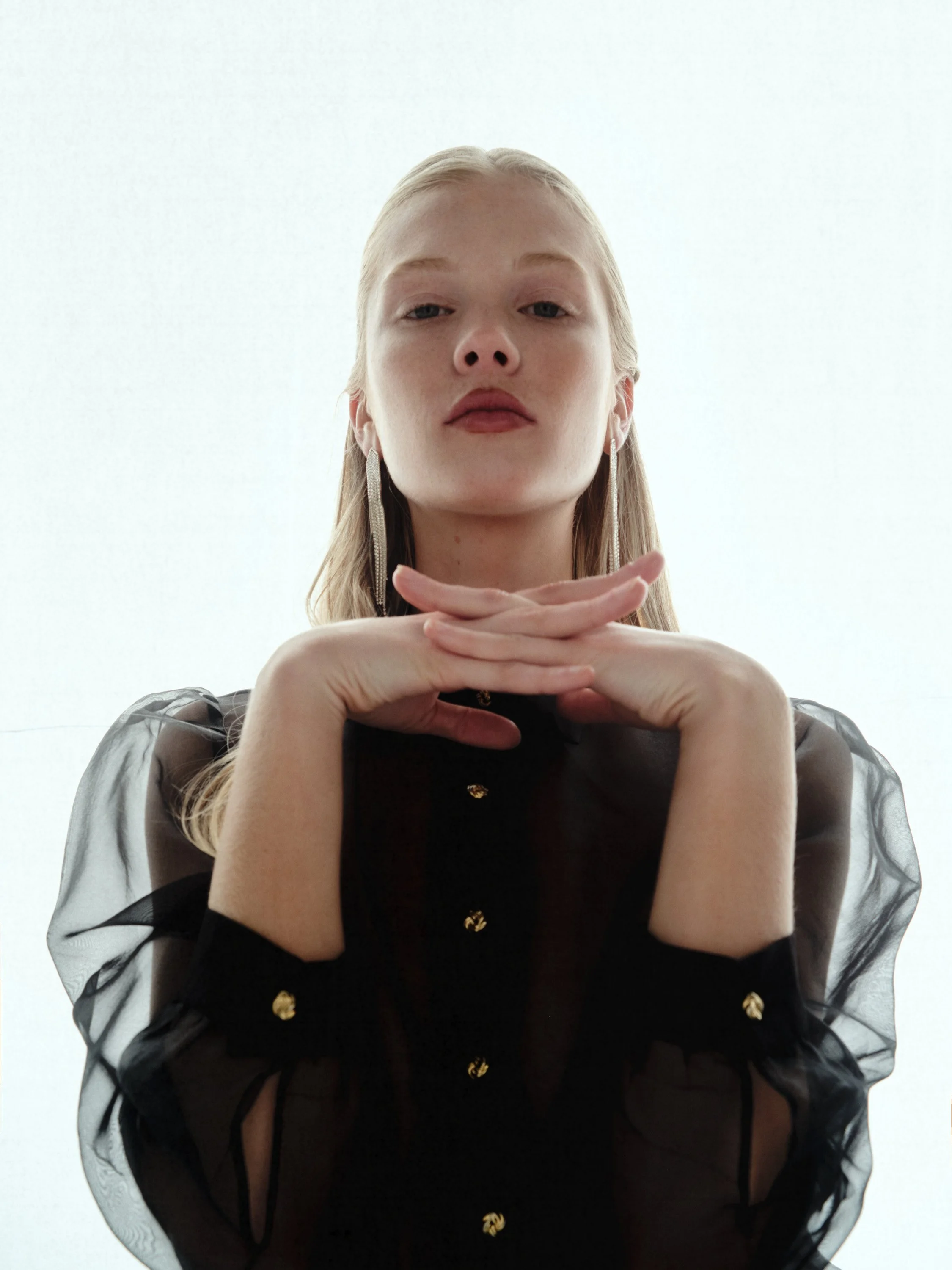 A woman with blonde hair, wearing long earrings, and a black sheer blouse with gold buttons, is looking down at the camera with her hands clasped under her chin, against a plain white background.