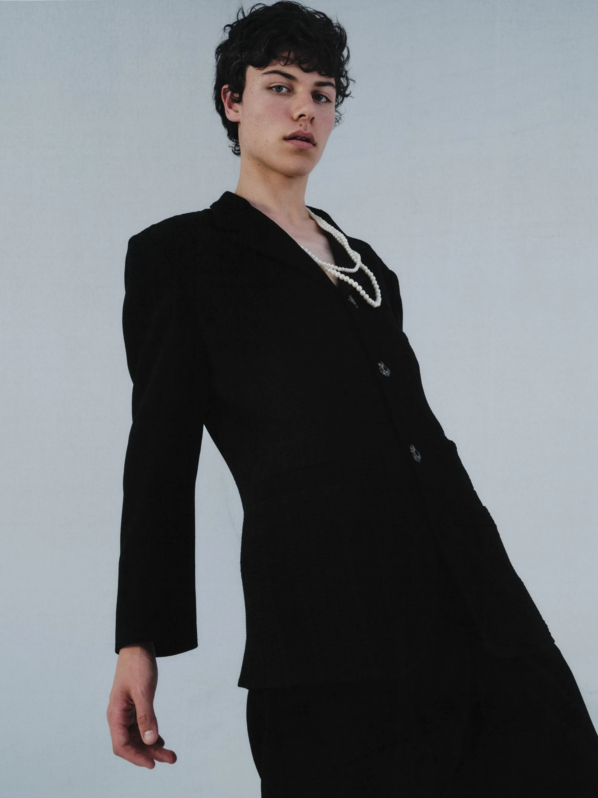 Young person with short, curly dark hair wearing a black blazer and a pearl necklace against a plain light background.