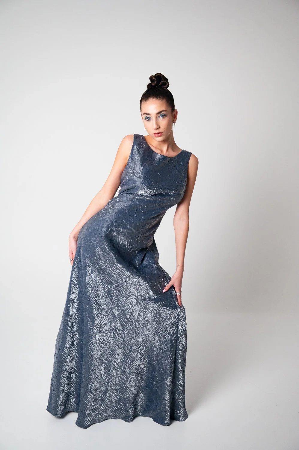 A woman wearing a long, shiny, gray evening gown with a textured pattern poses against a plain light gray background.