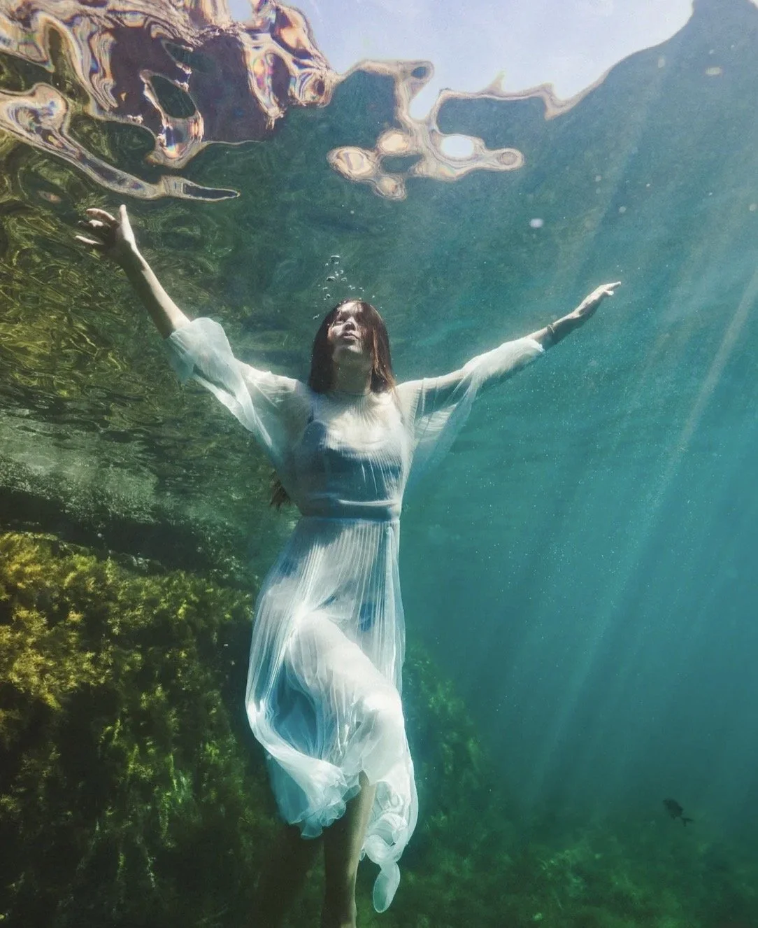Woman in a flowing white dress underwater with arms outstretched, sunlight filtering through water, green aquatic plants below, reflection of water's surface above.