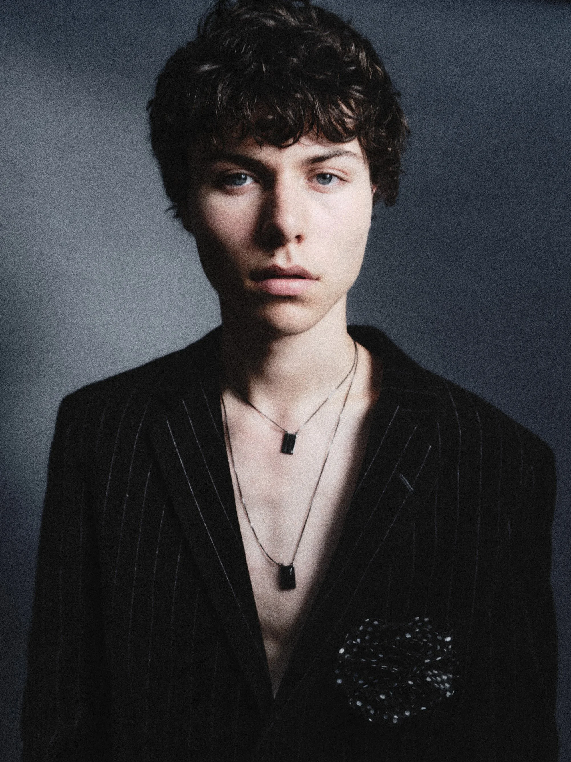 Portrait of a young woman with short, curly brown hair wearing a dark pinstripe blazer with a pocket square and layered necklaces against a dark background.