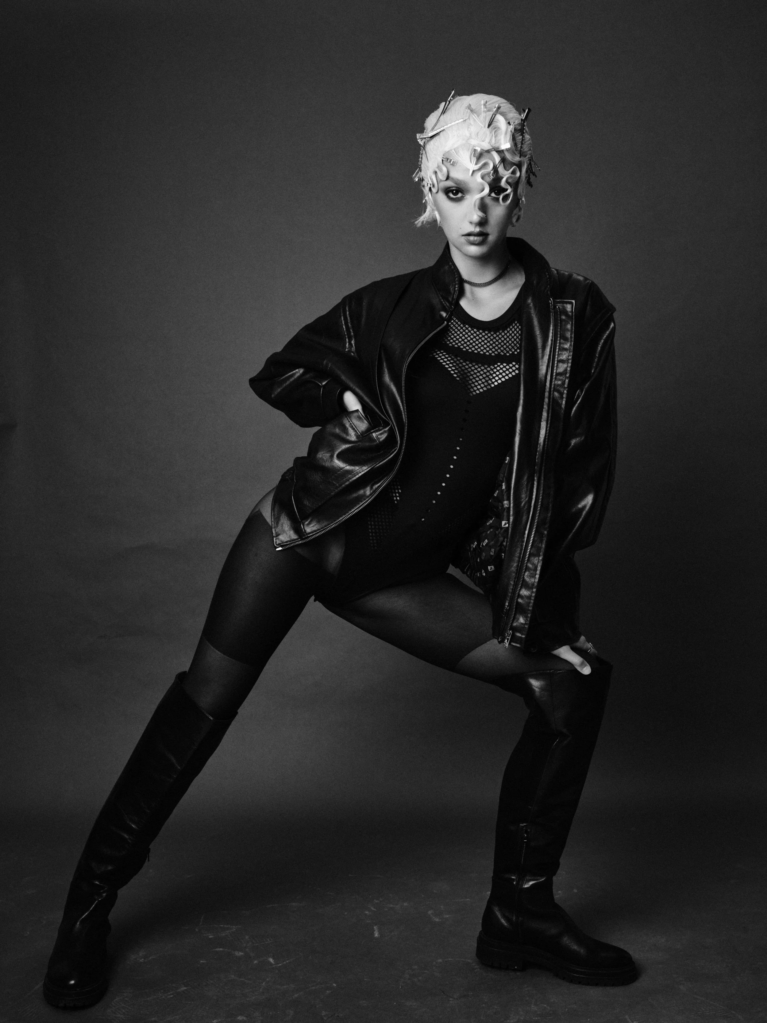 A woman with styled hair, wearing a leather jacket, fishnet top, shorts, tights, and knee-high boots, posing in a studio with a dark background.