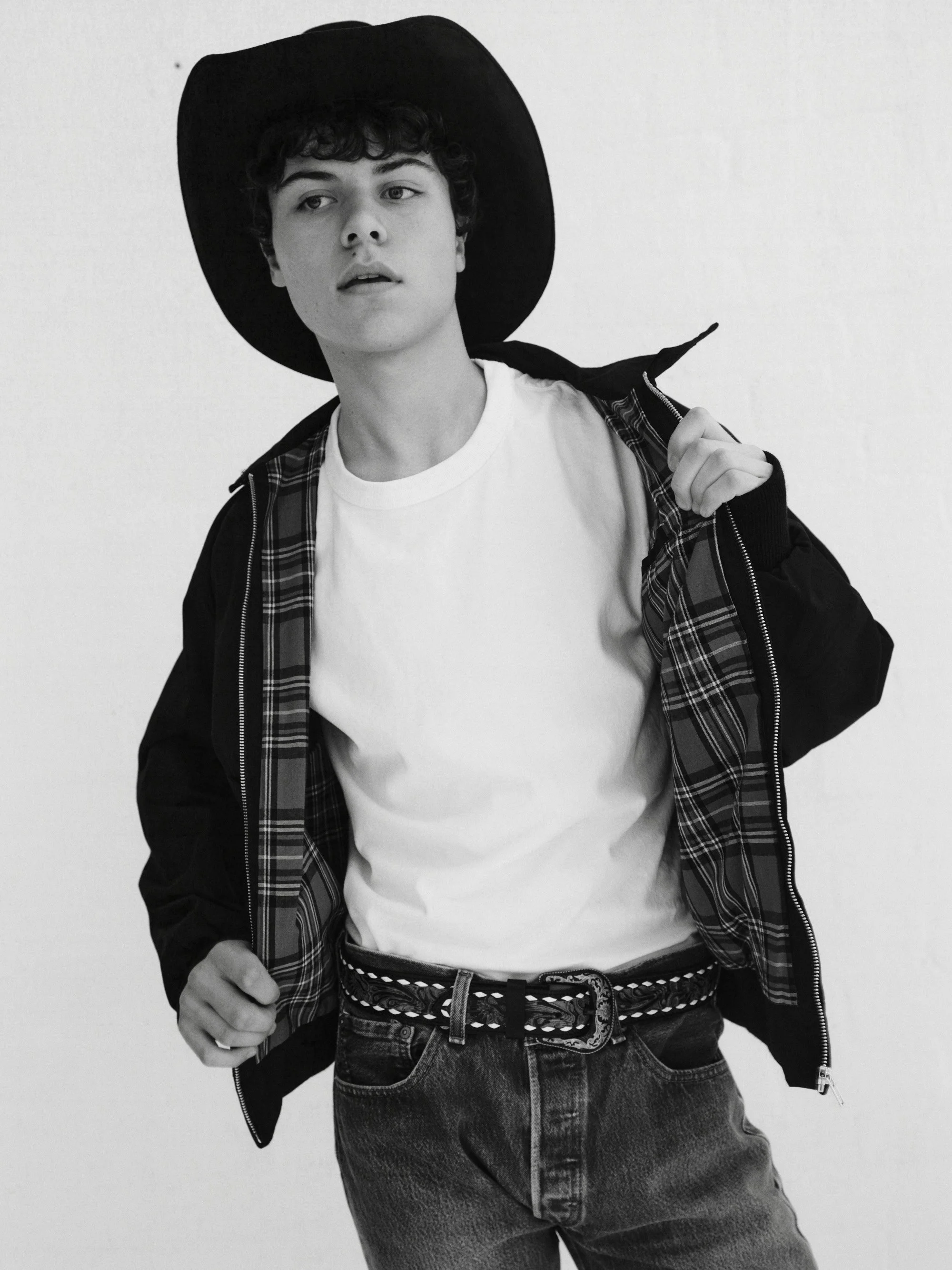Black and white photo of a young man wearing a large hat, white t-shirt, plaid jacket, and jeans, standing against a plain background.