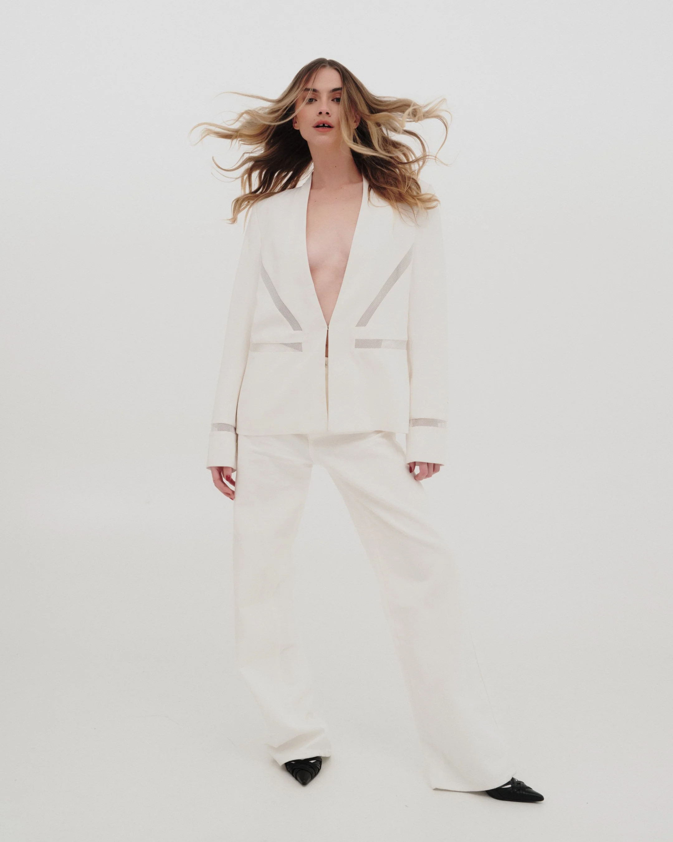 Woman with long, wavy hair wearing a white, tailored suit with a deep V neckline, standing against a plain white background.