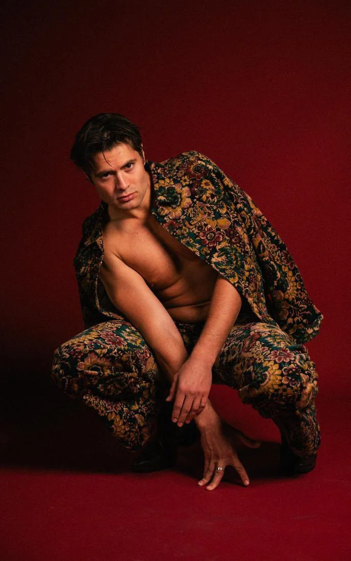 A young man with dark hair, dressed in a floral-patterned jacket and matching pants, crouching against a red backdrop, with one hand on the ground and the other resting on his knee, looking directly at the camera.