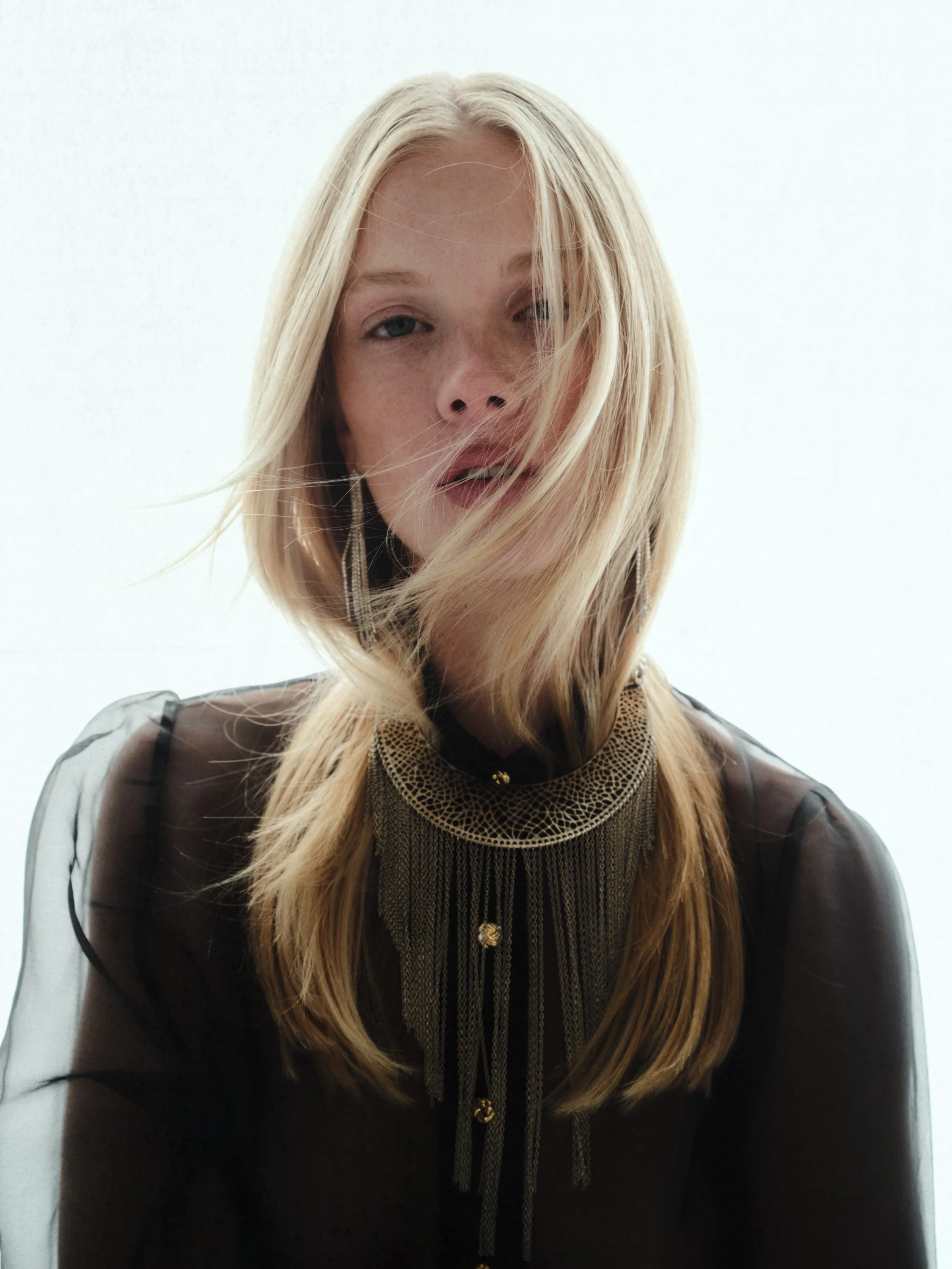A woman with blonde hair wearing a black sheer top and a gold jewelry necklace with chains and embellishments.