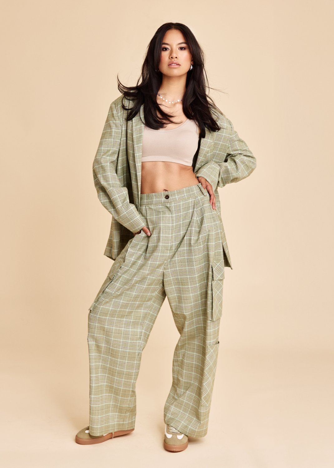 A young woman with long dark hair wearing a light beige tank top, a plaid blazer, matching plaid wide-leg pants, and sneakers, standing with one hand on her hip against a neutral background.