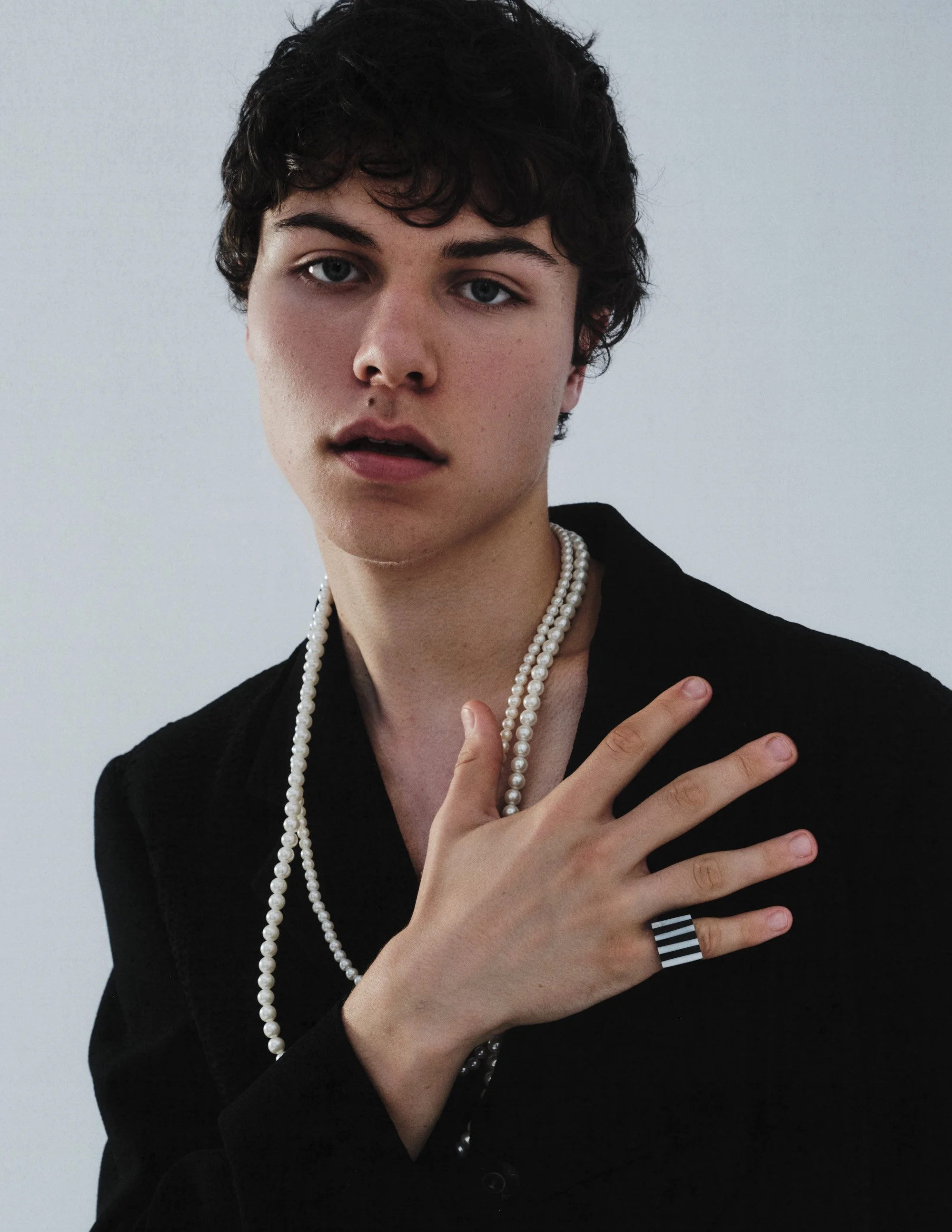 A young person with dark curly hair, wearing a black blazer, multiple pearl necklaces, and a black and white striped ring, posing against a plain white background.