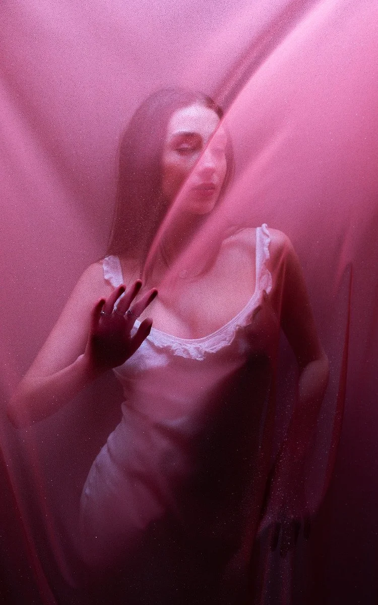 A woman in a satin nightgown standing behind a pink, semi-transparent curtain, touching it with her hand. The scene has a pinkish hue and a dreamy atmosphere.