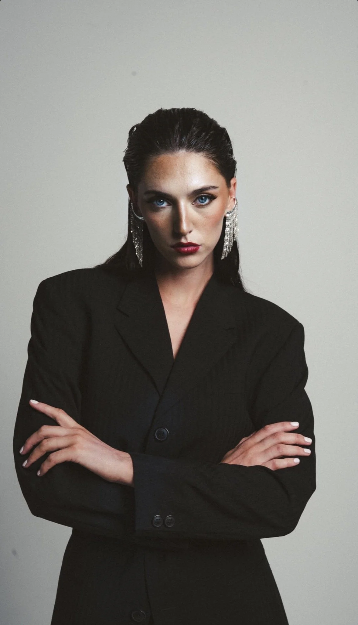 A woman with slicked-back dark hair, blue eyes, and red lipstick, wearing a black suit and large sparkly earrings, posing with crossed arms against a plain light-colored background.