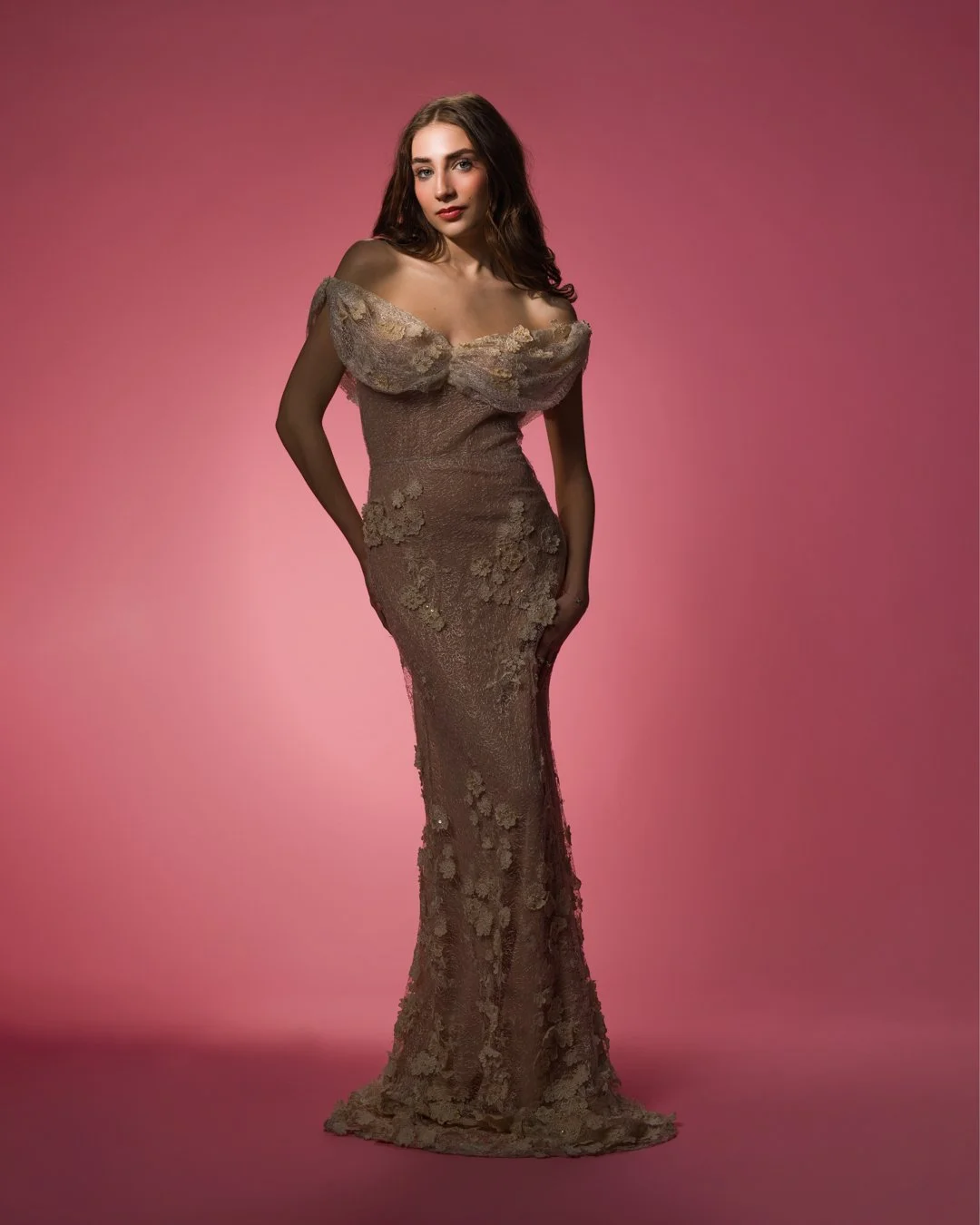 Woman in a gold lace, off-shoulder gown with floral details, standing against pink background.