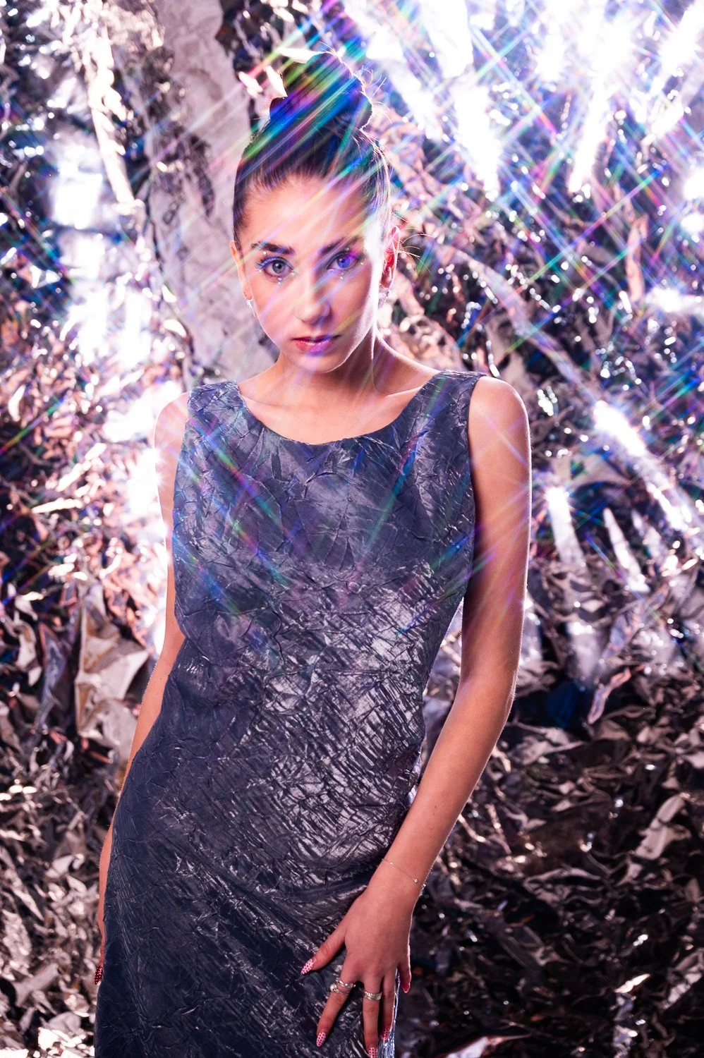 A woman with her hair in a bun, wearing a shiny, textured black dress, standing in front of crumpled metallic foil background with rainbow light streaks.
