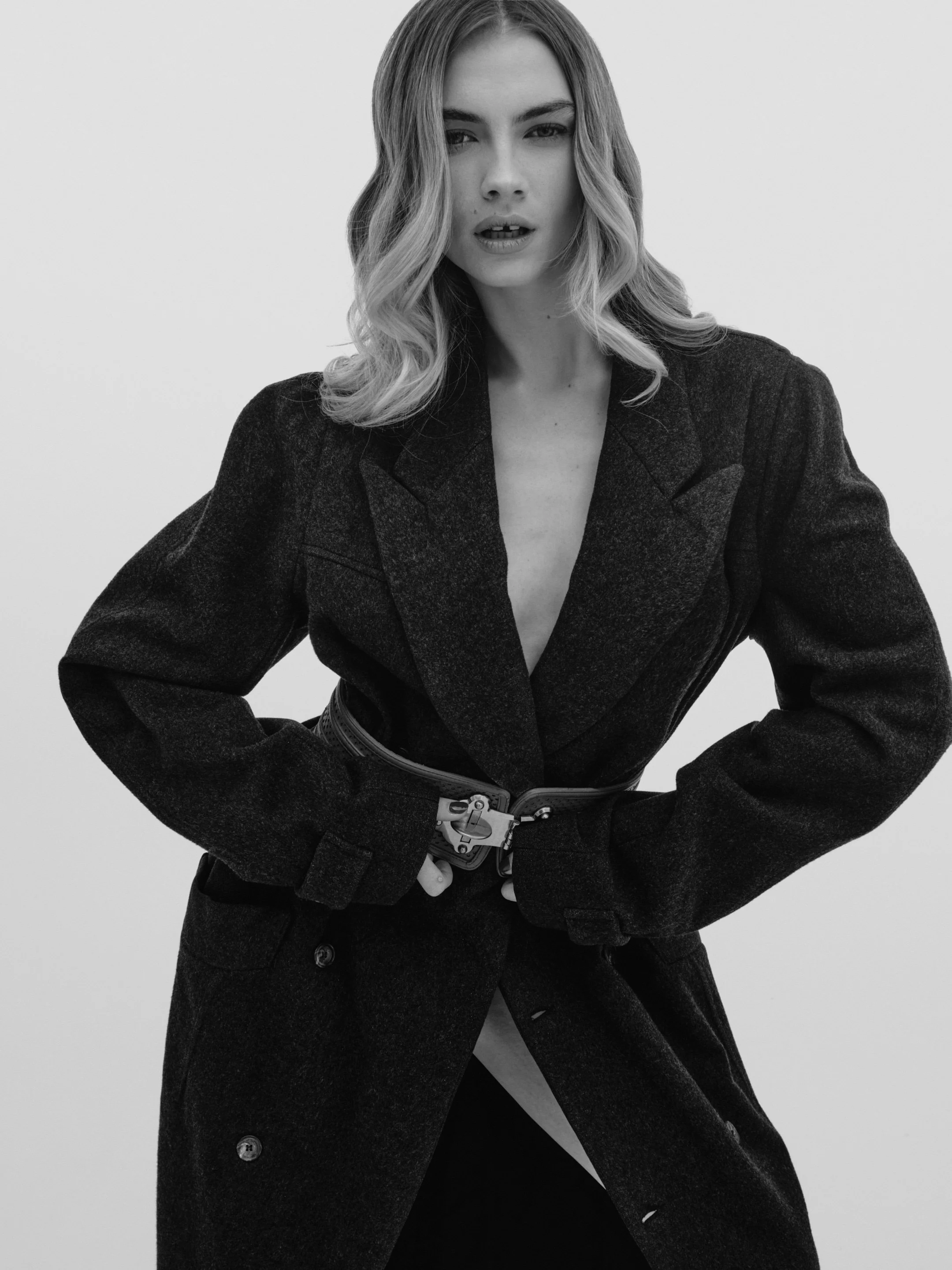 Black and white photo of a woman with wavy hair, wearing a dark blazer and a wide belt, standing with hands on hips against a plain background.