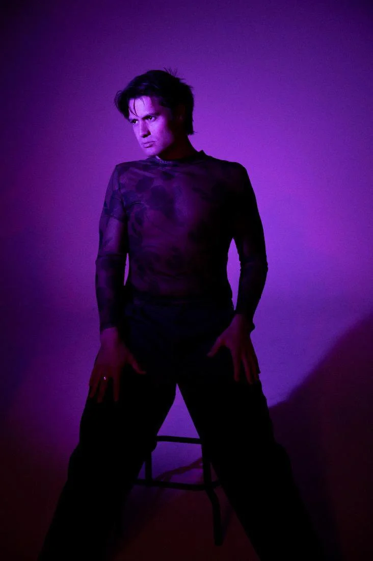 Person with short dark hair wearing a sheer, long-sleeved purple top, posing with hands on thighs against a purple-lit background.