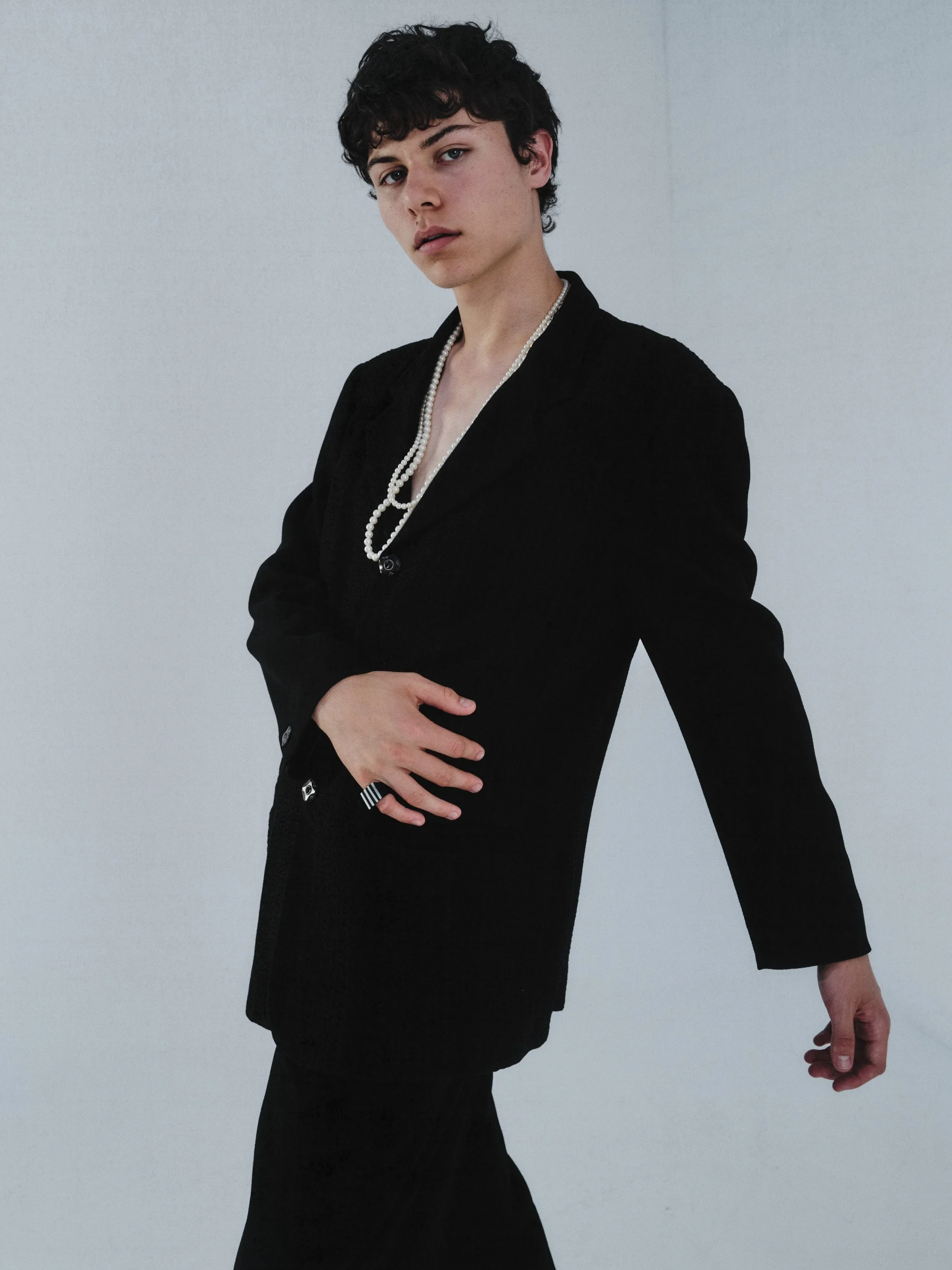 A young person with short curly dark hair, wearing a black suit and a pearl necklace, standing against a plain light gray background.