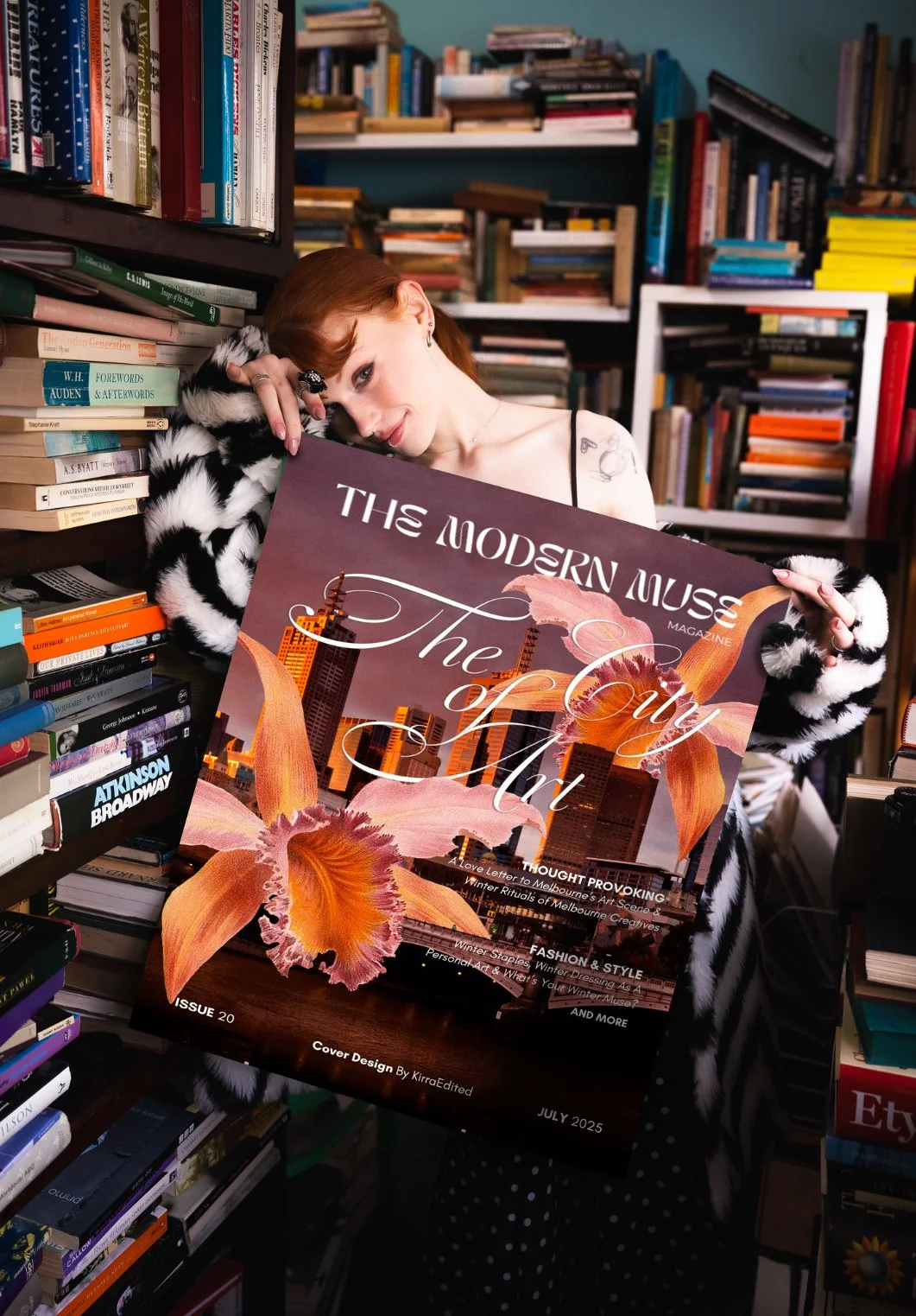 A woman with red hair, dressed in a black and white fur coat, holding a magazine titled 'The Modern Muse' in a bookstore or library surrounded by shelves filled with books.