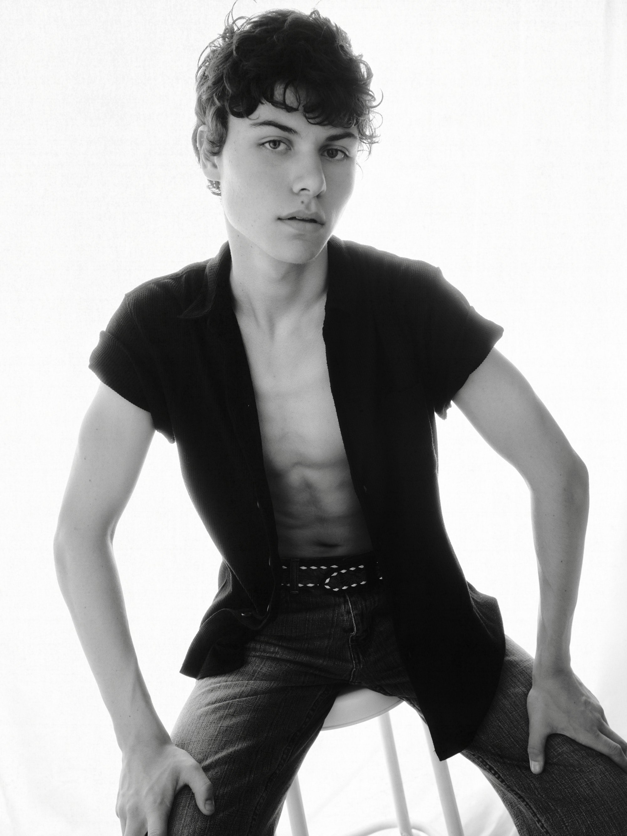 A black-and-white photo of a young person with short, curly hair sitting on a stool, wearing an open shirt and jeans, looking directly at the camera with a serious expression.