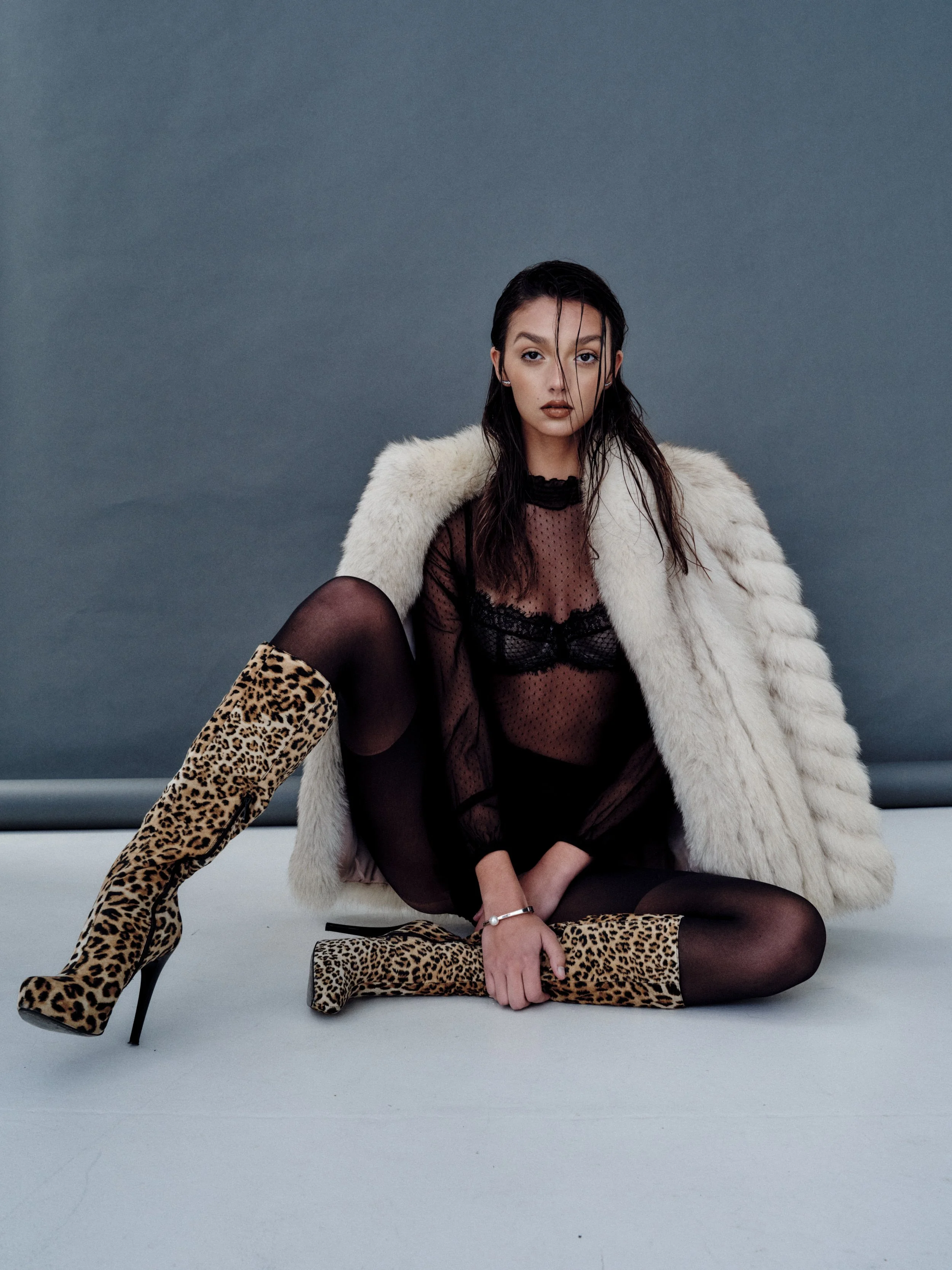 Woman sitting on a white floor, wearing leopard print high-heeled boots, black sheer clothing, and a white fur coat, with a grey background.