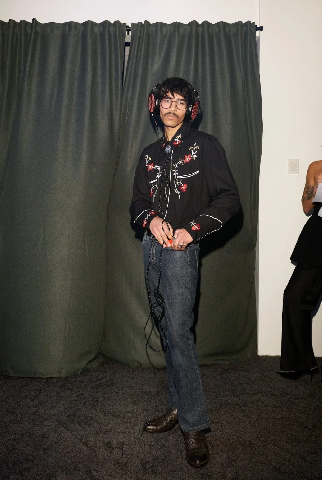 A young man wearing glasses, a black embroidered Western-style shirt, jeans, and brown boots, standing in front of dark curtains, holding headphones around his neck.