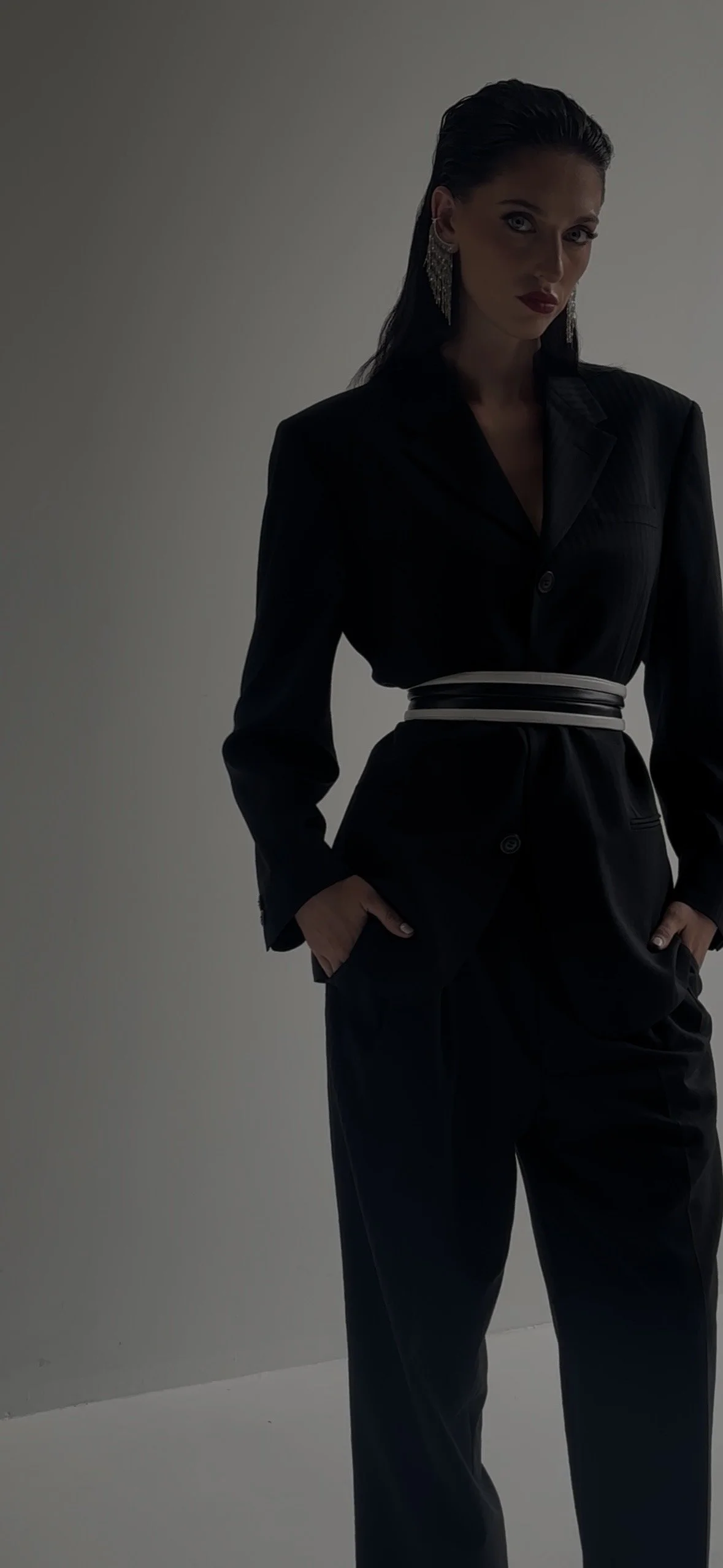 Woman in black suit with hands in pockets, wearing large earrings, standing against a plain background.