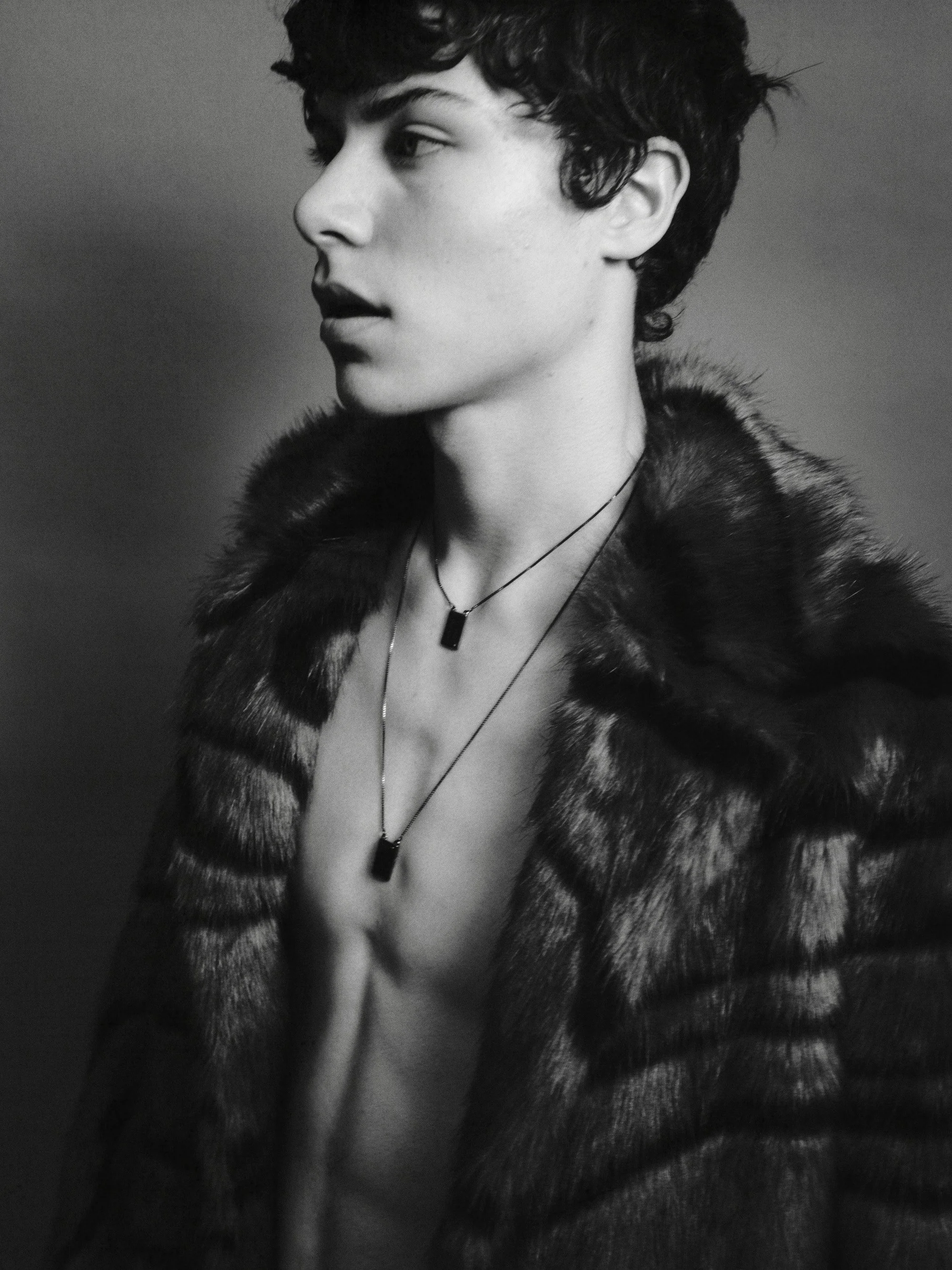 A black-and-white portrait of a young person with short, curly hair, wearing a fur coat and layered necklaces, looking to the side.