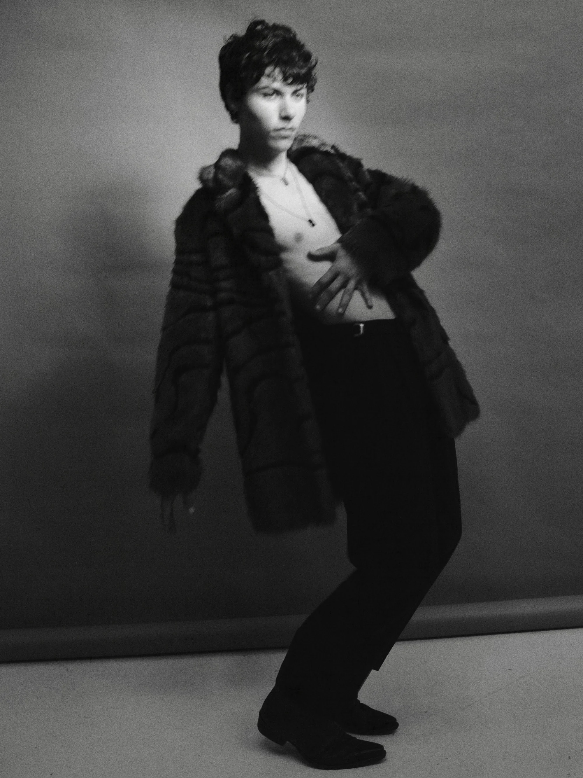 Black and white photo of a person with short curly hair wearing a fur coat, a white top, black pants, and shoes, standing against a plain background.