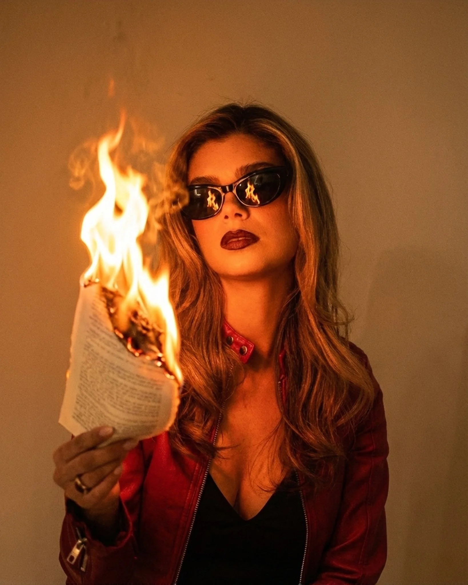 A woman with brown hair, wearing dark sunglasses and a red leather jacket, holding a burning book.
