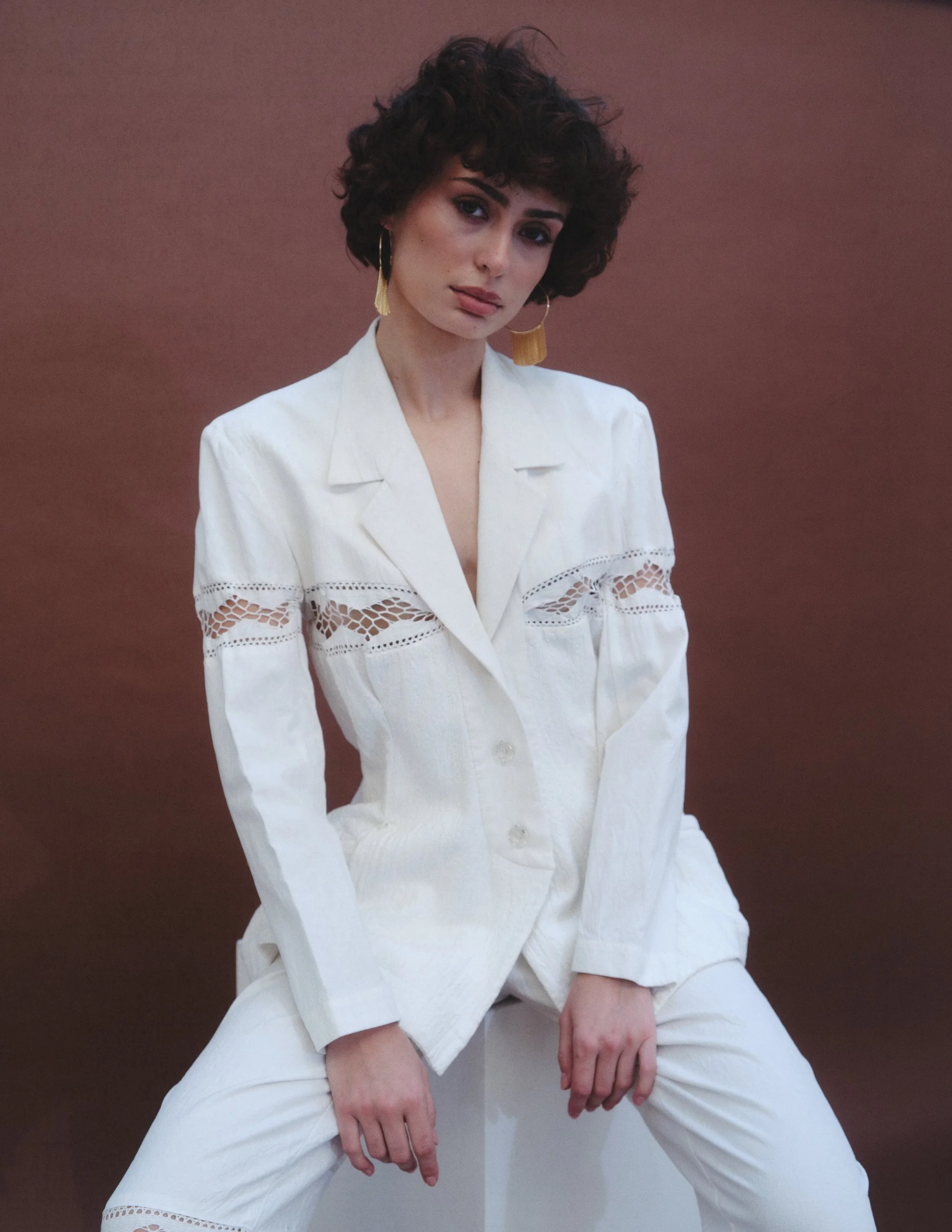 Woman with short curly hair wearing a white blazer with lace details and matching white pants, sitting on a white block against a brown background.