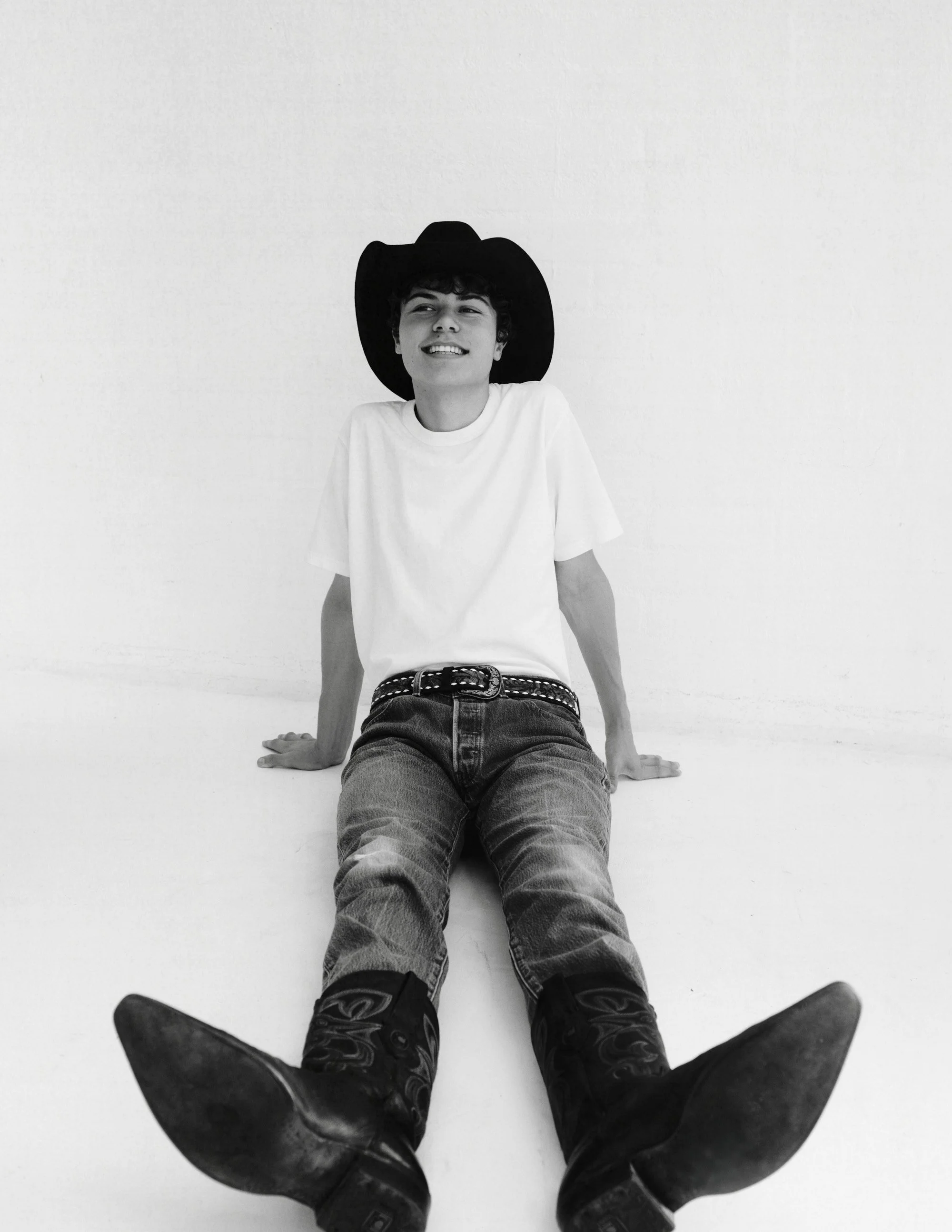 A young man wearing a cowboy hat, white t-shirt, jeans, and cowboy boots sitting on the floor leaning back with a relaxed smile.