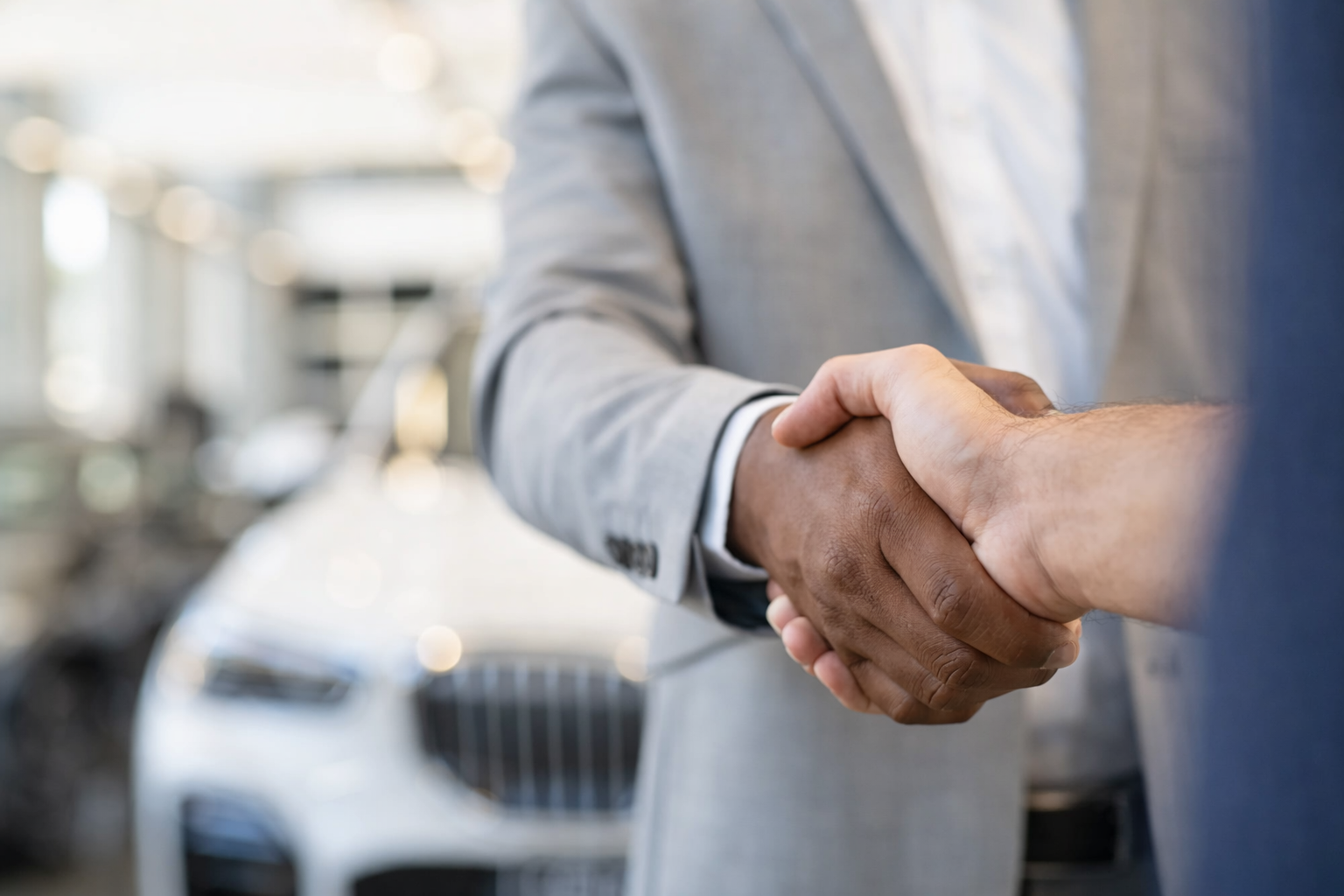 Close-up of business professionals shaking hands in a car dealership showroom, with cars in the background.