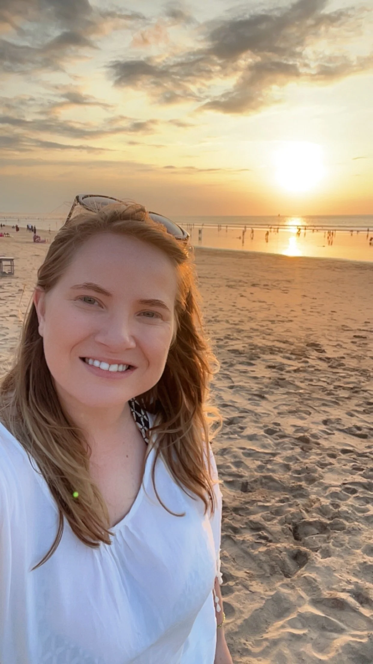 A woman taking a selfie on the beach during sunset, with the ocean, sky with clouds, and people in the background.