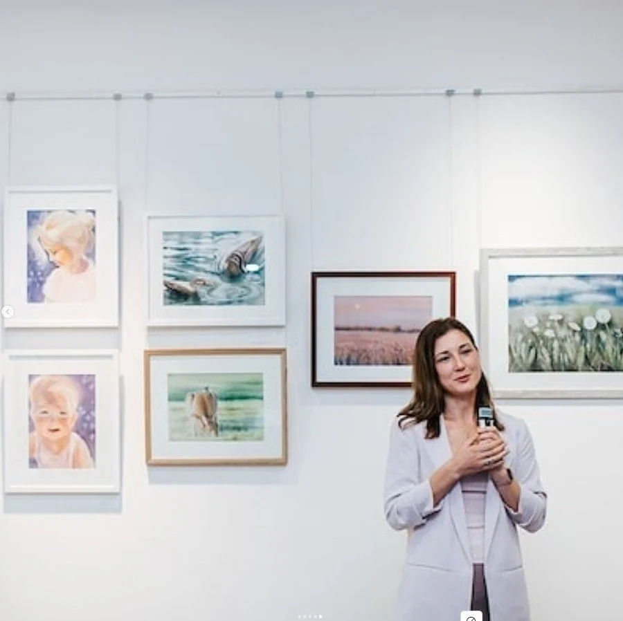 A woman in a white blazer holding a smartphone in an art gallery with various framed photographs and paintings on the white wall behind her.