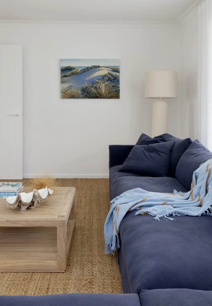 A cozy living room with a navy blue sofa, a light wooden coffee table holding books and a decorative bowl, a white floor lamp, a landscape painting of dunes and grasses on the wall, and sunlight coming through a curtain.