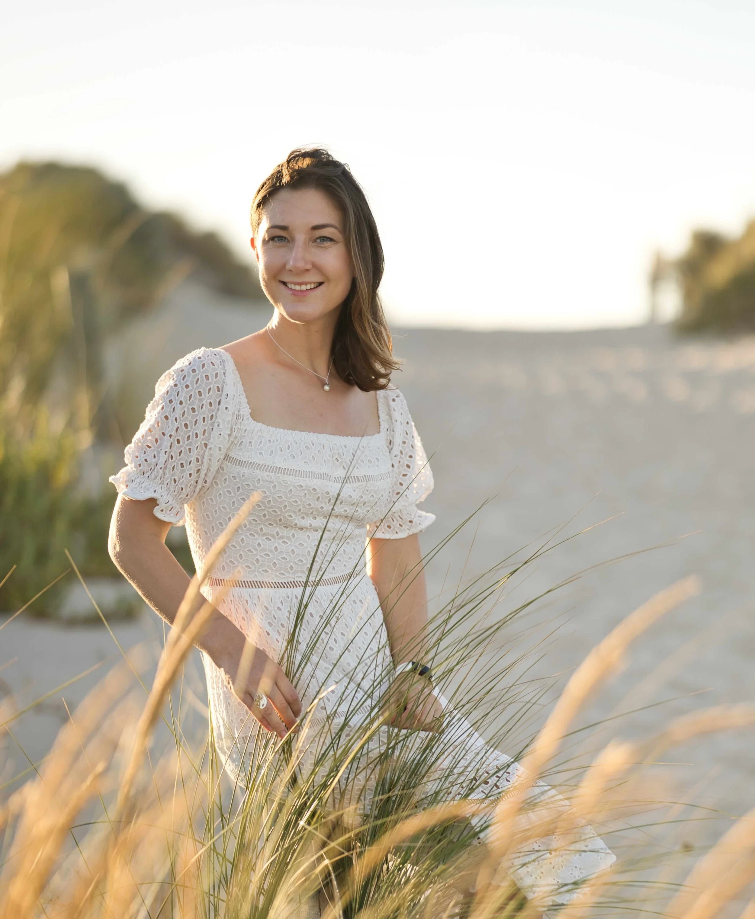 Woman in a white dress sitting on a sandy beach with grass, smiling, during sunset.