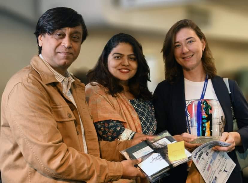 Three people standing together at an indoor event, holding brochures and smiling at the camera.