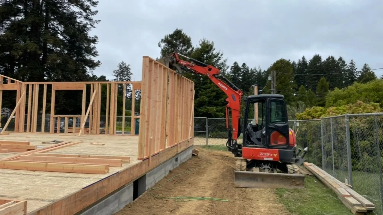 A small red excavator is building the wooden frame of a house on a construction site, with a chain-link fence and trees in the background.