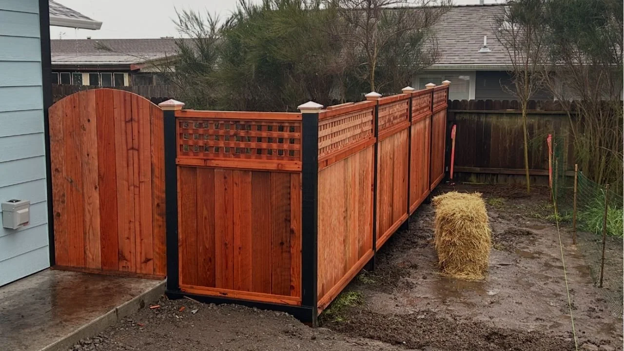 A newly installed wooden privacy fence with black posts and decorative lattice panels along the top, enclosing a backyard. There is a wooden gate at one end, a bale of hay on muddy ground, and a few trees and neighboring houses in the background.