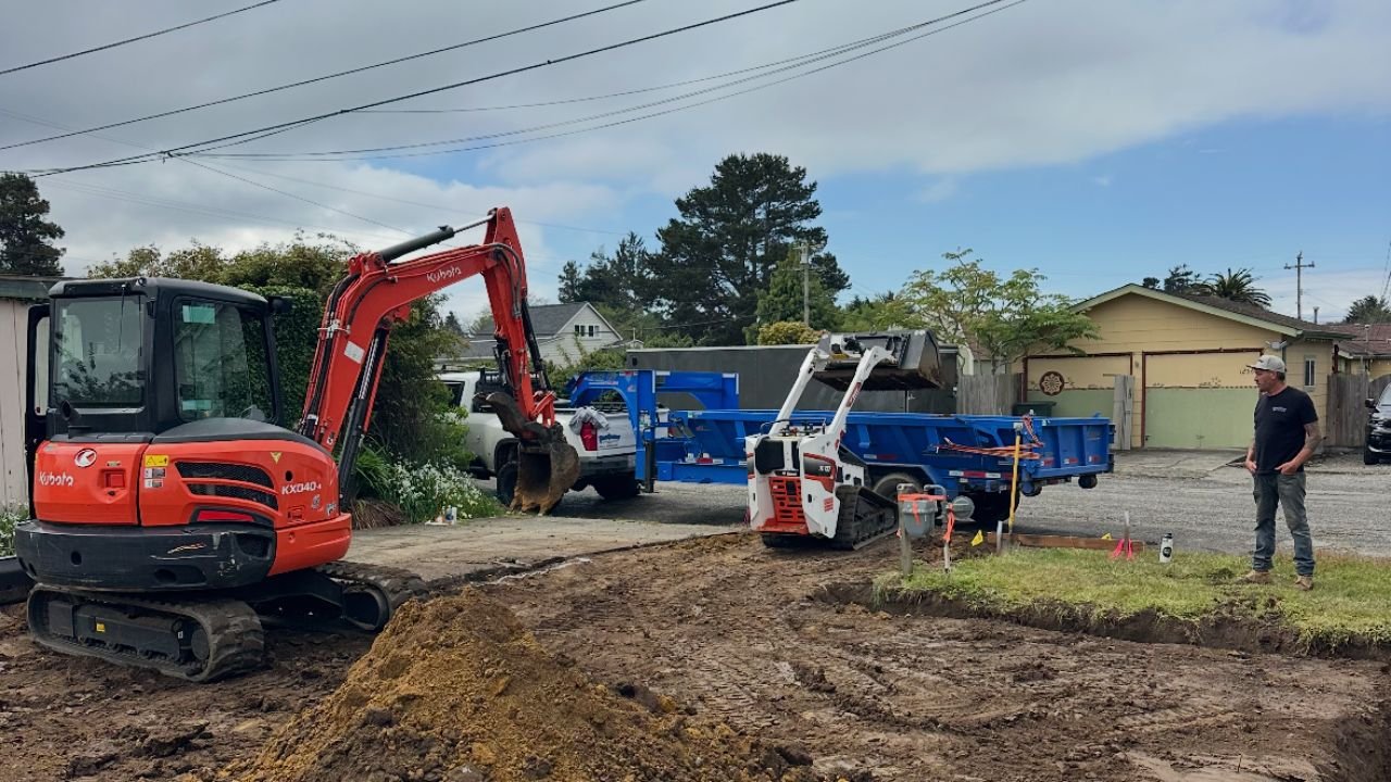 Construction site with an orange excavator, a small white and orange skid-steer loader, and a blue dump truck on a partly cloudy day. A man in a black shirt and gray cap observes the work, which involves digging and earthmoving.