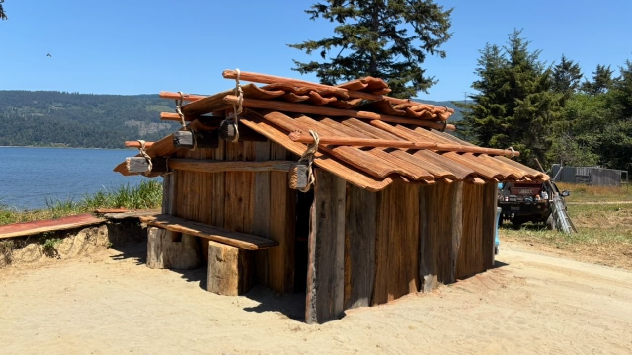 A small wooden structure with a roof made of wooden tiles, situated on sandy ground near a body of water, with trees in the background.